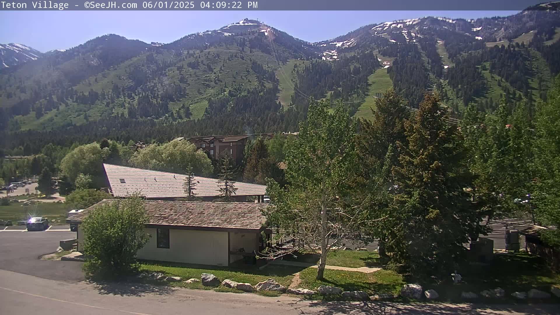 A sunny day reveals a mountain range with patches of snow, overlooking a village with buildings and trees.