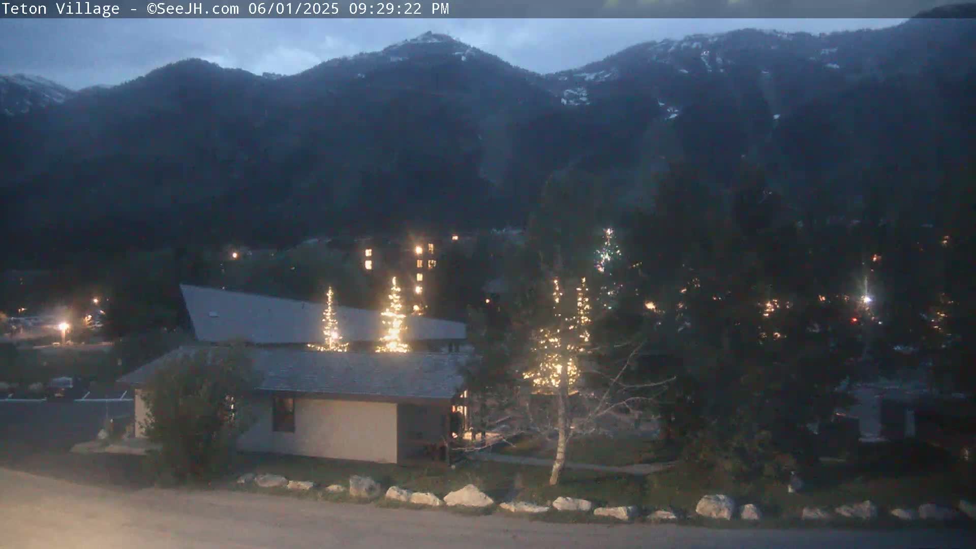 A low-light outdoor scene shows a building with illuminated trees in front, a parking lot, and mountains in the background under a twilight sky.
