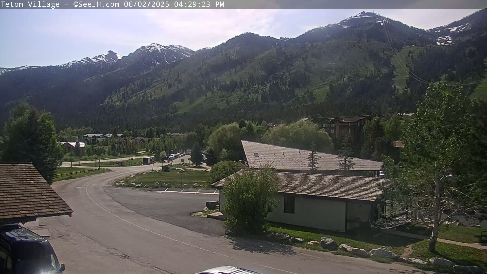 A sunny view of a mountain range with some snow on the peaks, overlooking a village with buildings and a road curving through a grassy area.