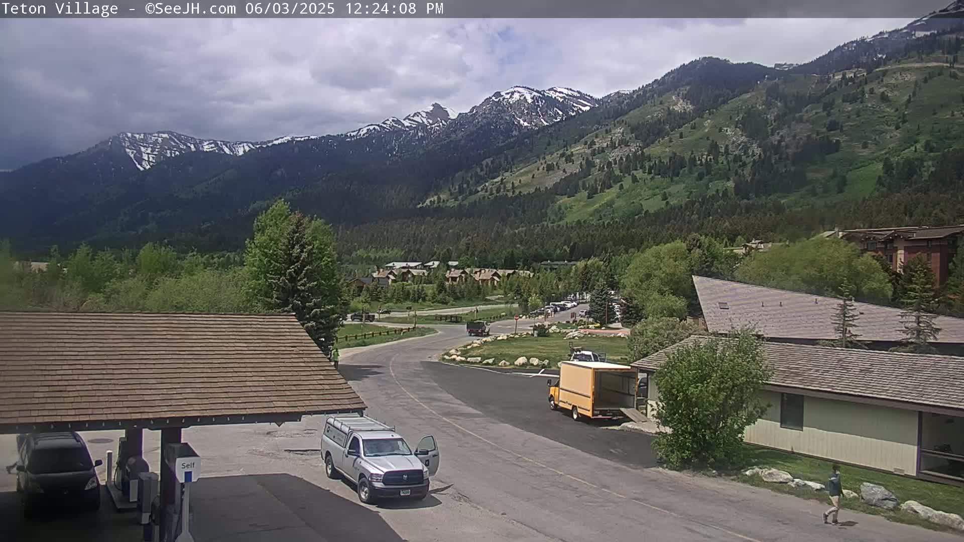 A partly cloudy day shows a mountain valley with snow-capped peaks in the background, a road with several vehicles, and buildings in the foreground.