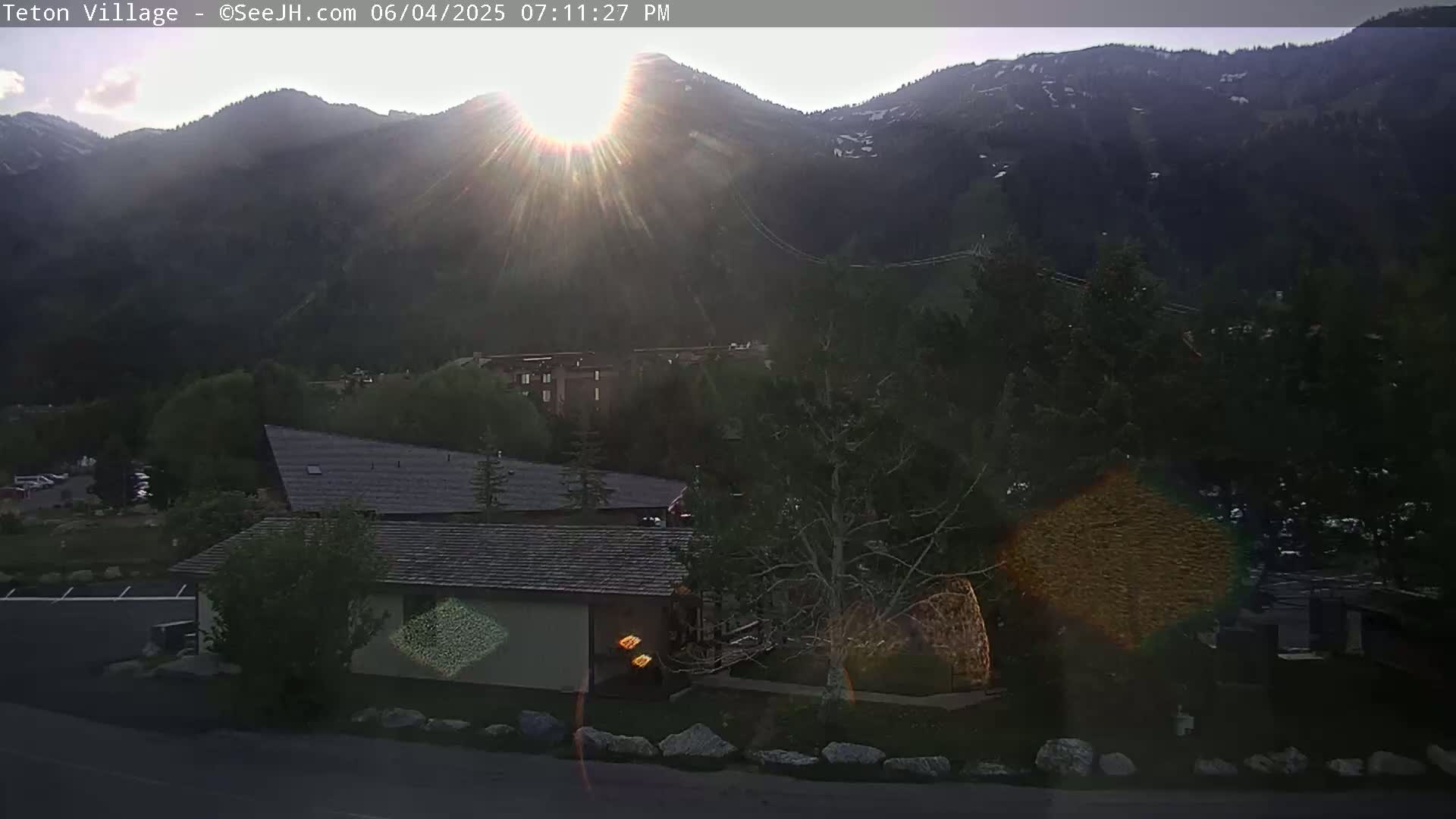 A low-angle, sunset view shows a mountain range in the background, with a building and trees in the foreground under a mostly clear sky.