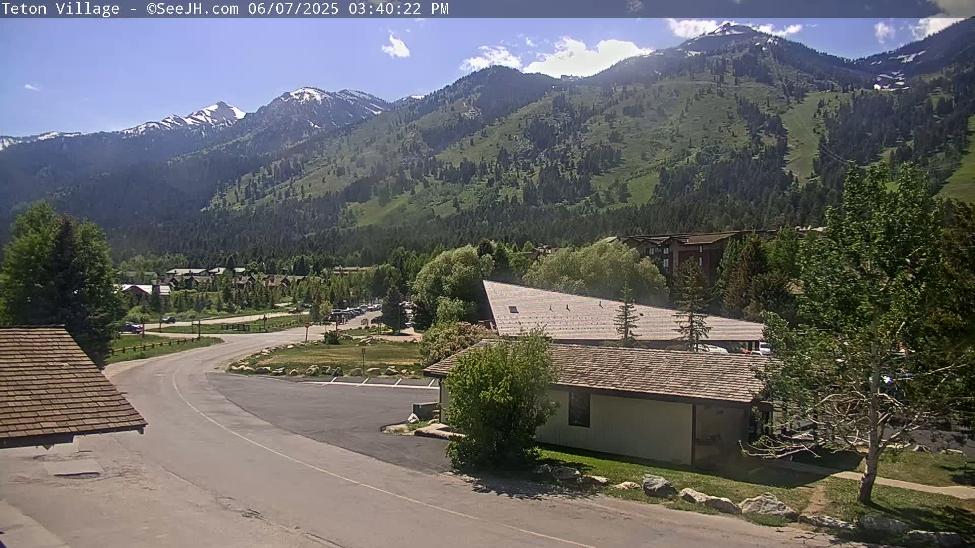 A sunny day reveals a mountain range partially covered in snow, overlooking a village with roads, buildings, and lush green trees.