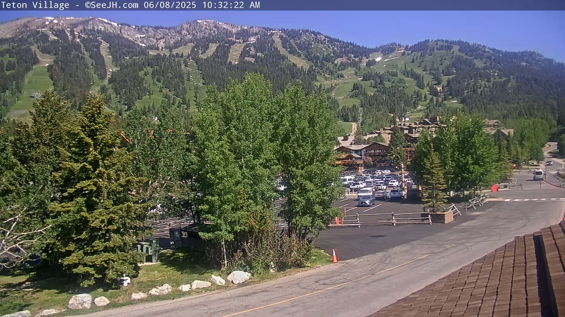A sunny view of a mountainside ski resort with a parking lot and buildings, surrounded by green trees.