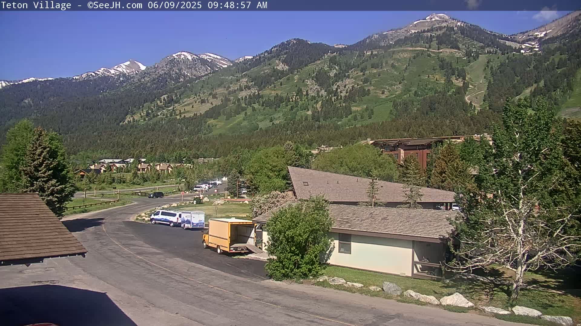 A sunny view of a mountainside town with snow-capped peaks in the background, featuring a parking lot with several vehicles and a low-slung building.