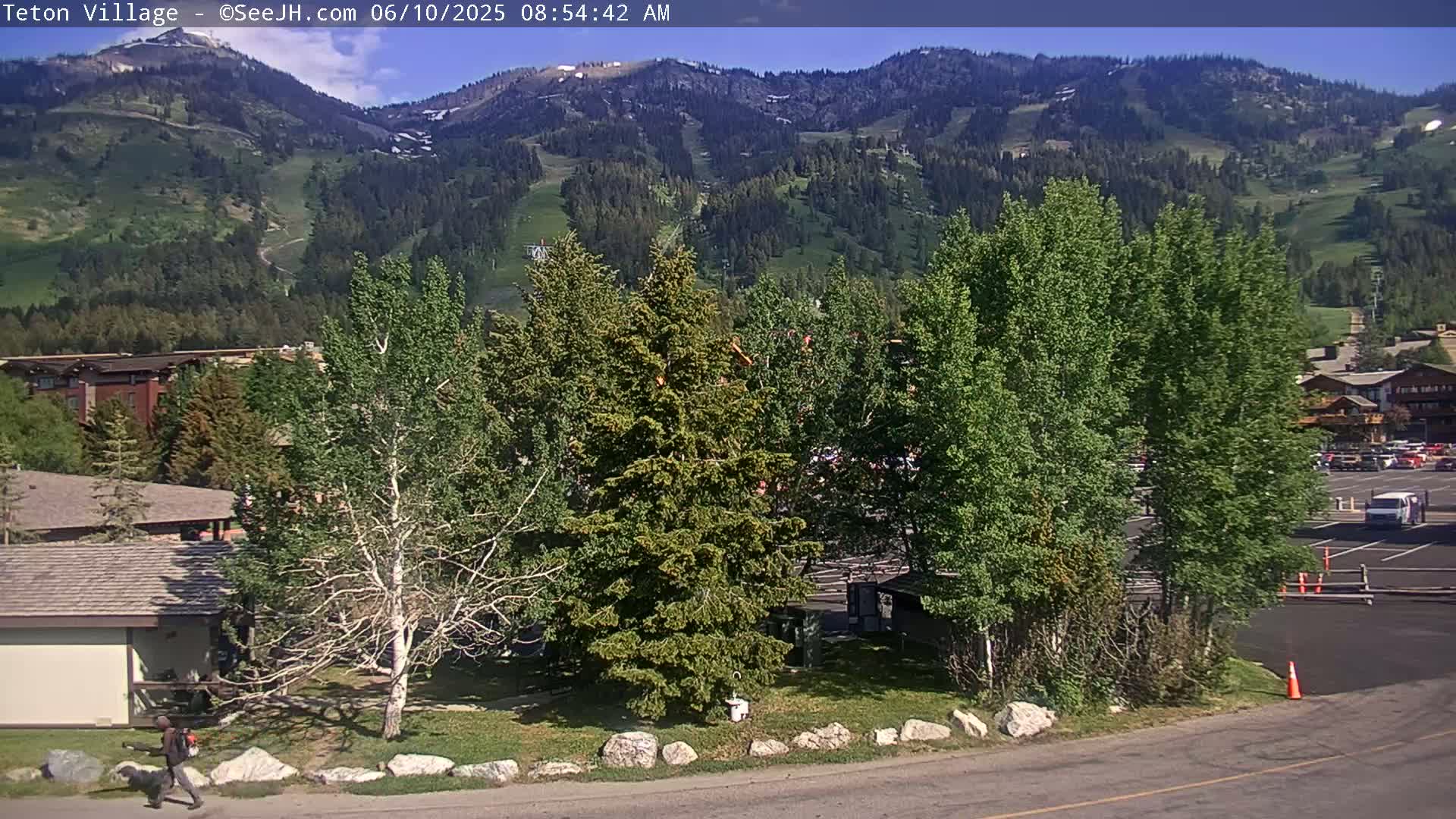 A sunny view of a grassy area with several evergreen and deciduous trees, a parking lot, and buildings, all nestled at the base of a mountain range with patches of snow.