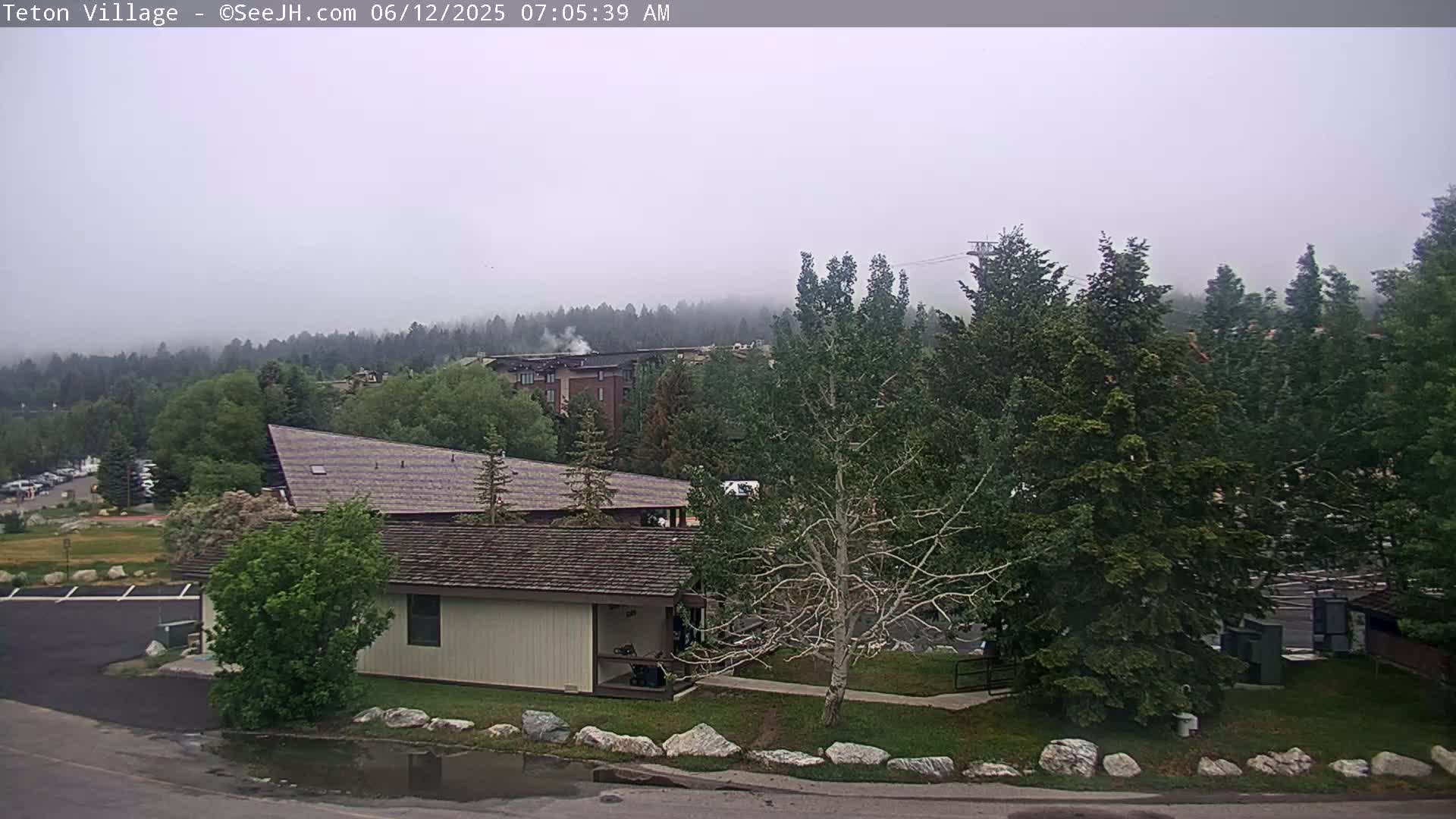 A foggy morning scene shows a low building with a peaked roof, surrounded by trees and a parking lot, with a mountain range and other buildings visible in the distance.