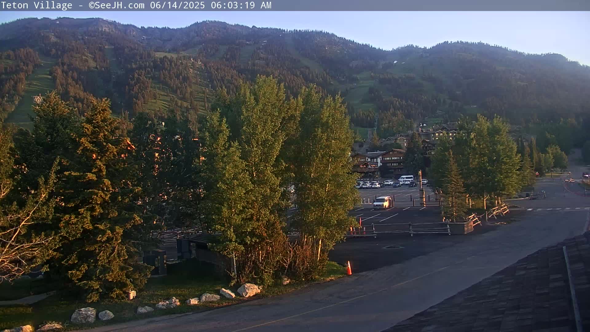 A parking lot and several buildings are situated in the foreground of a mountainside village under a clear sky at sunrise.