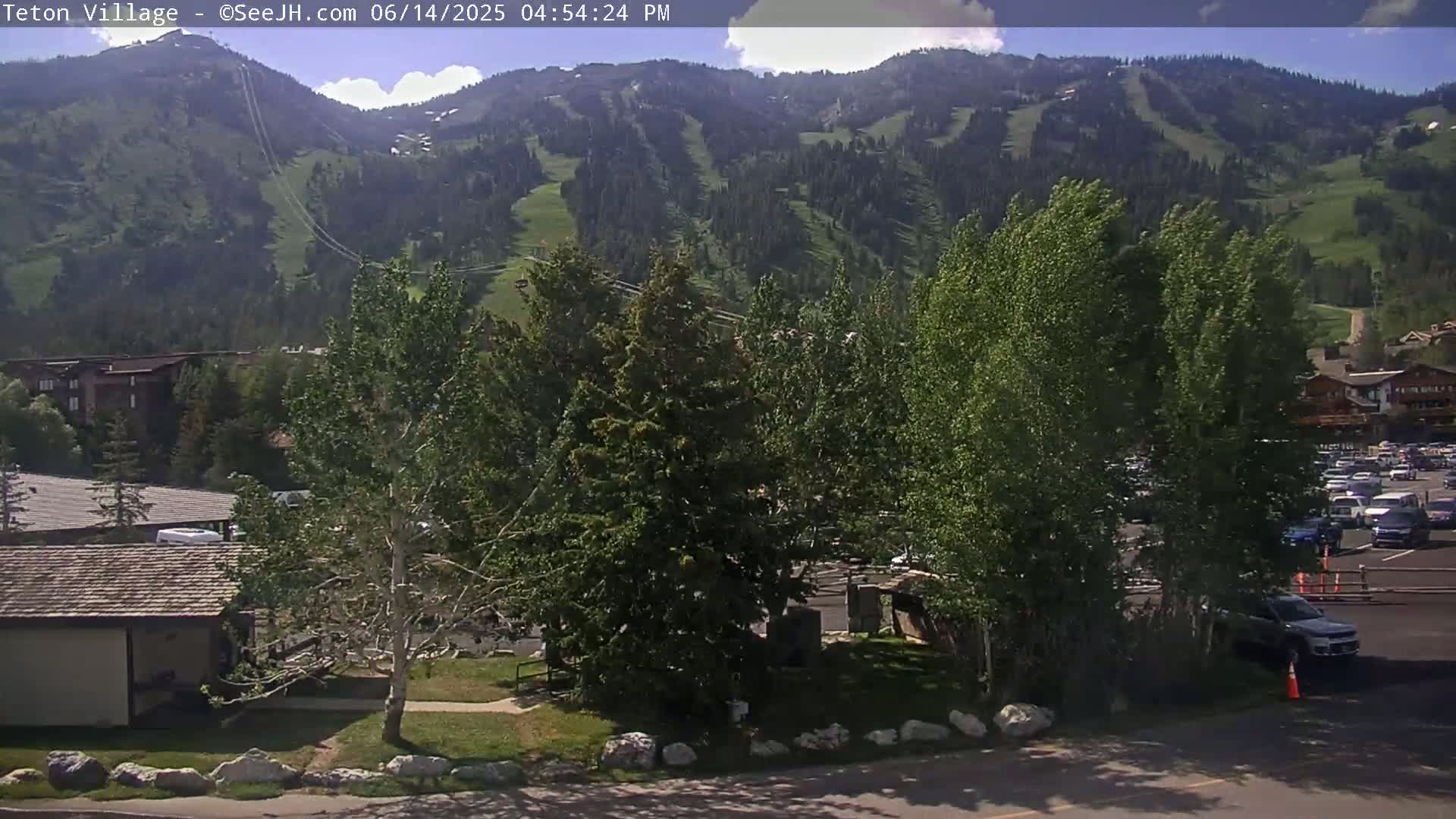 A sunny view of a village nestled at the base of a mountain range, featuring green trees, buildings, and a parking lot.