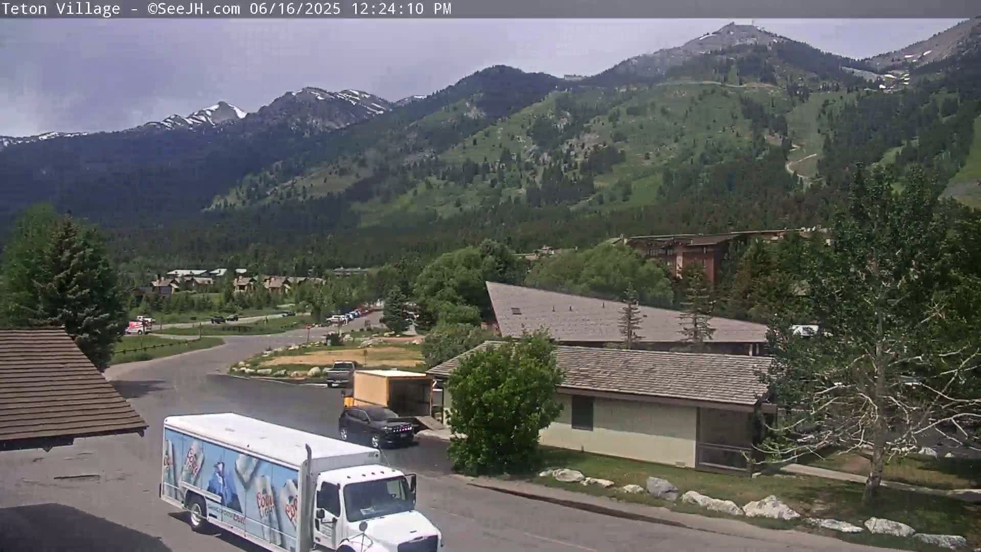 A partly cloudy day reveals a mountain vista overlooking a parking lot with a delivery truck and several buildings;  greenery blankets the slopes.