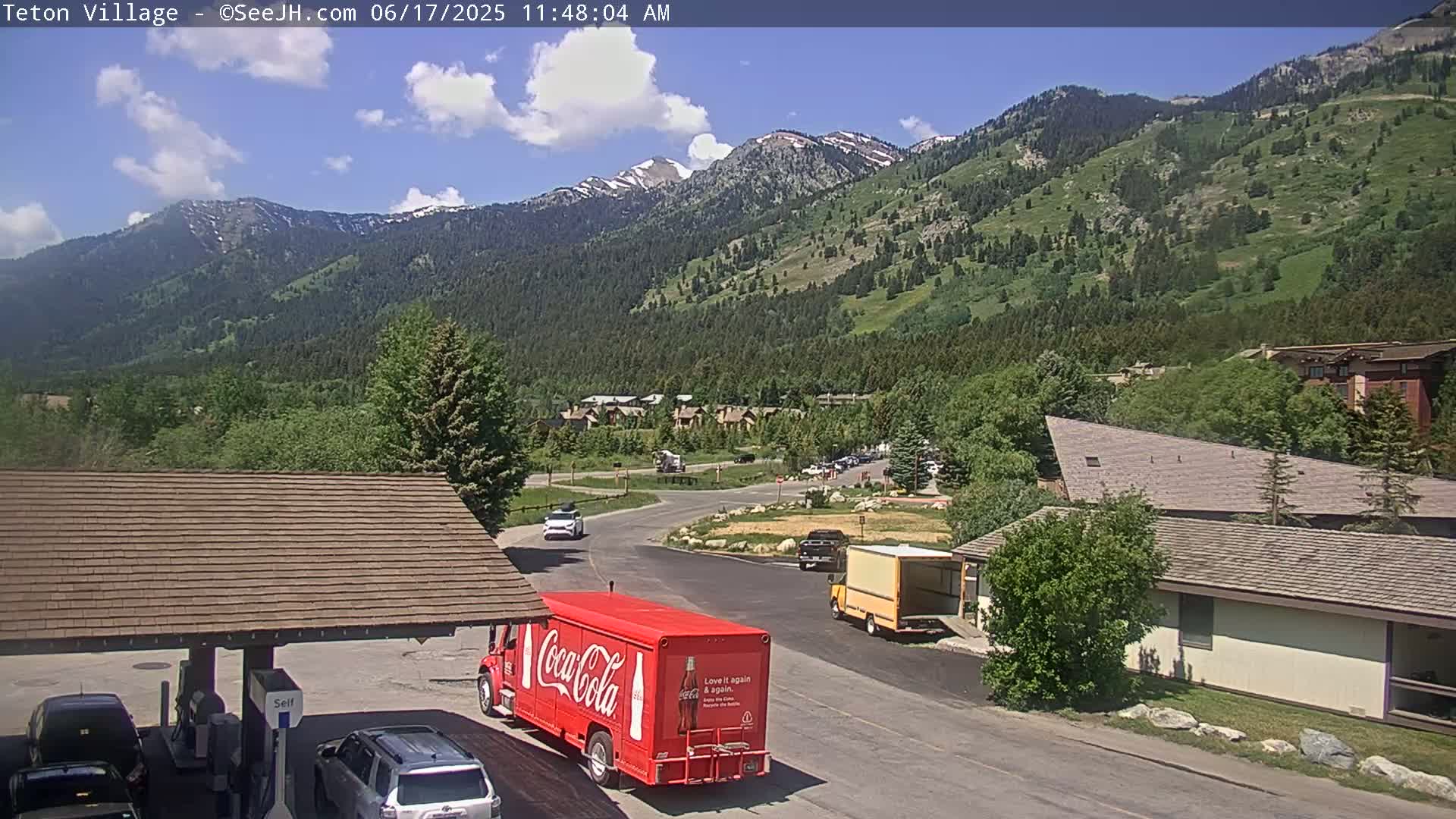 A partly cloudy day shows a mountain village with a Coca-Cola truck parked near a gas station, with mountains and trees in the background.