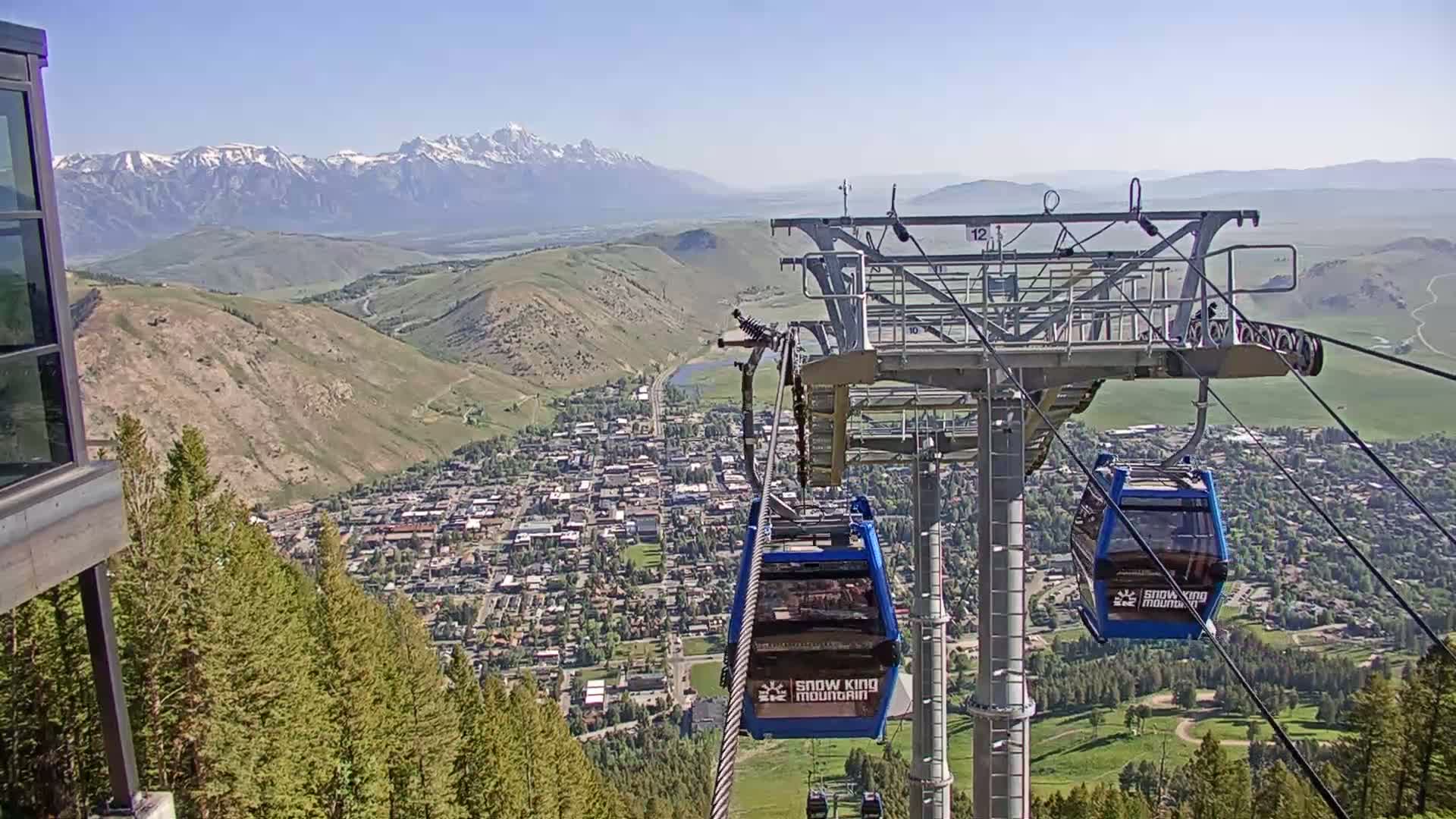 A chairlift ascends a mountain overlooking a town nestled in a valley, with snow-capped mountains in the background under a clear, sunny sky.