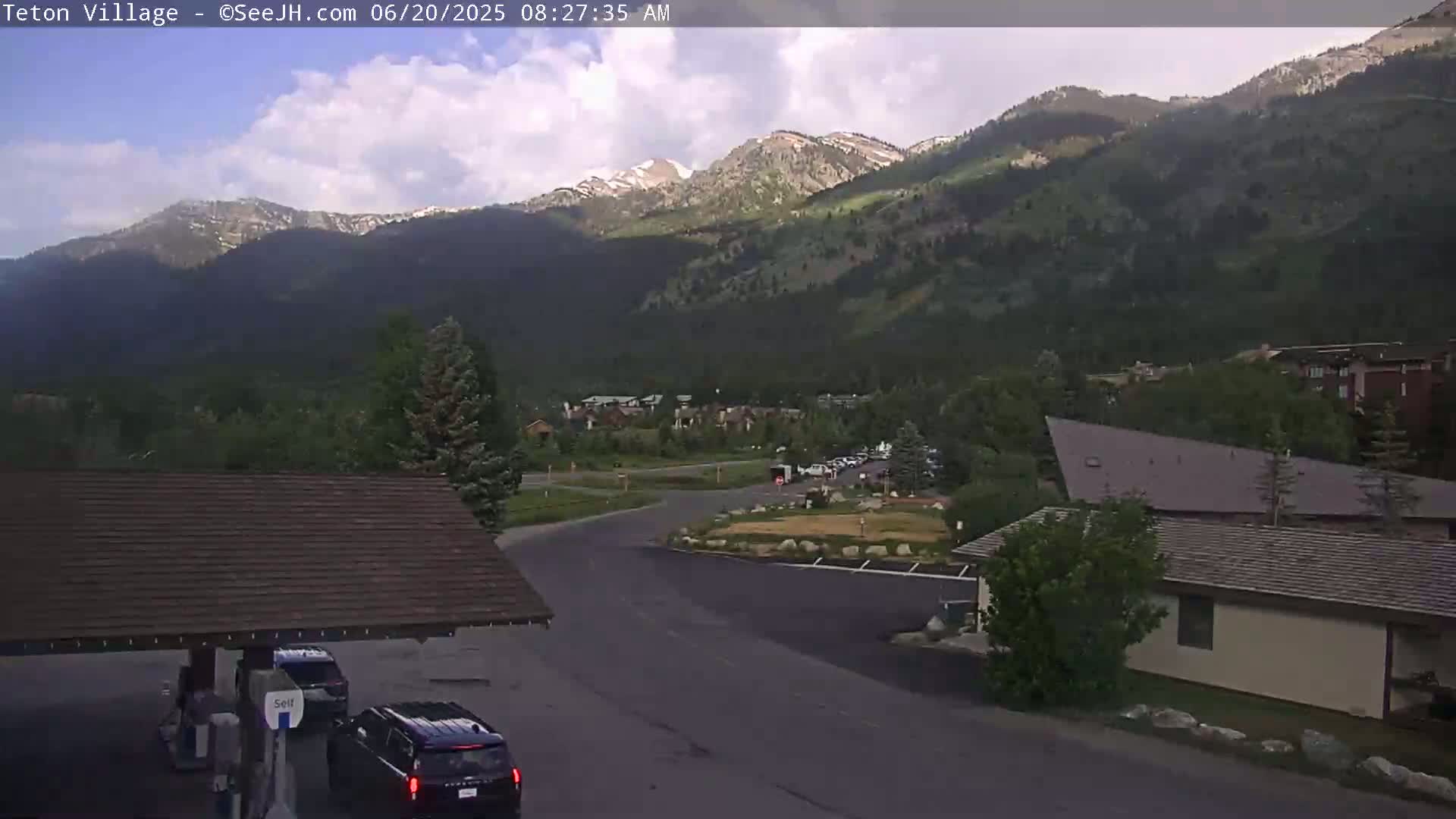 A partly cloudy morning scene shows a mountain village nestled in a valley, with a gas station and several vehicles visible in the foreground.