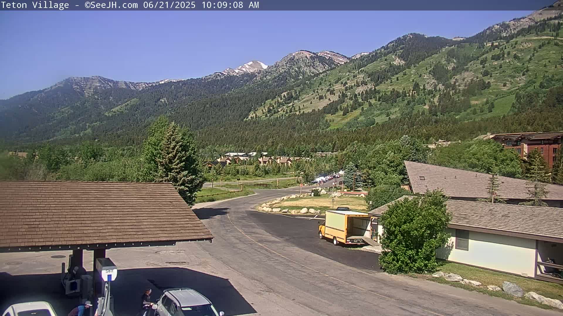 A sunny mountain village scene shows a roadway curving past buildings, a gas station, and a yellow delivery truck, with snow-capped mountains in the background.