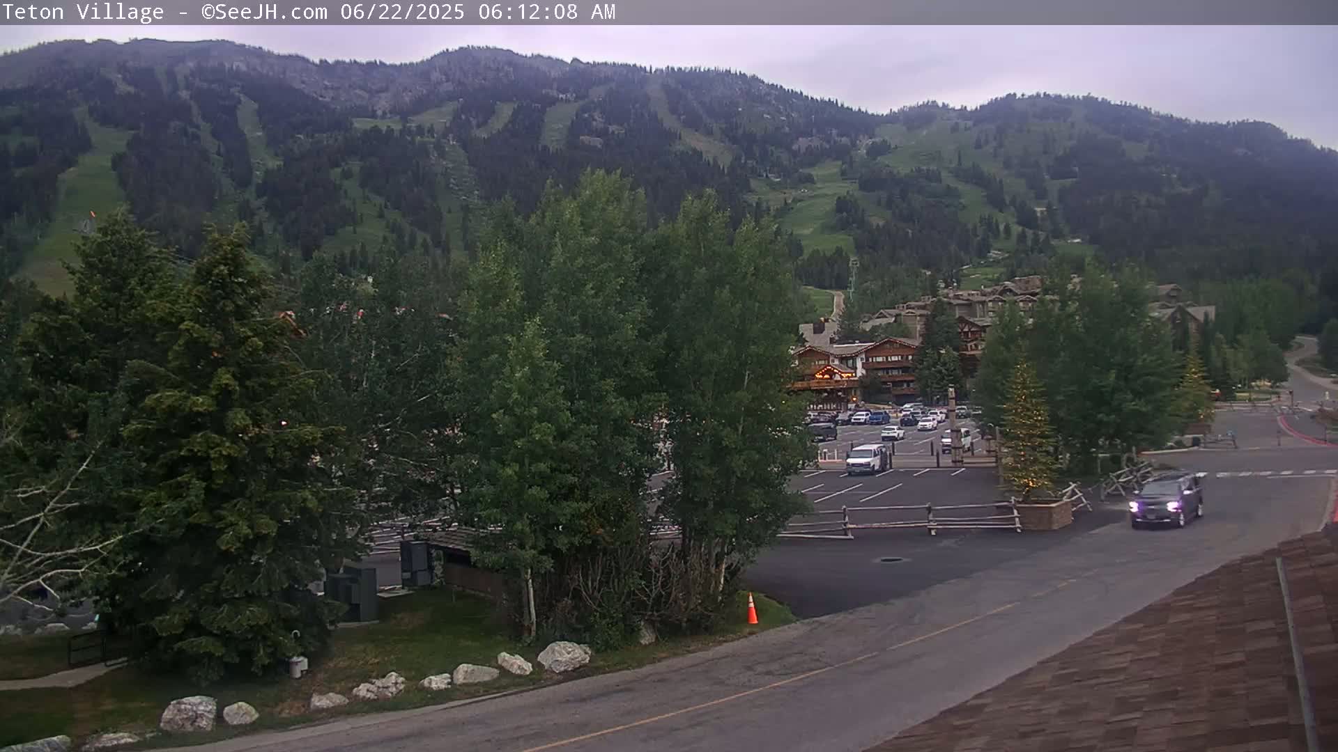 A mostly cloudy day reveals a mountainside with ski runs, a parking lot with several vehicles, a road, and some buildings near lush green trees.