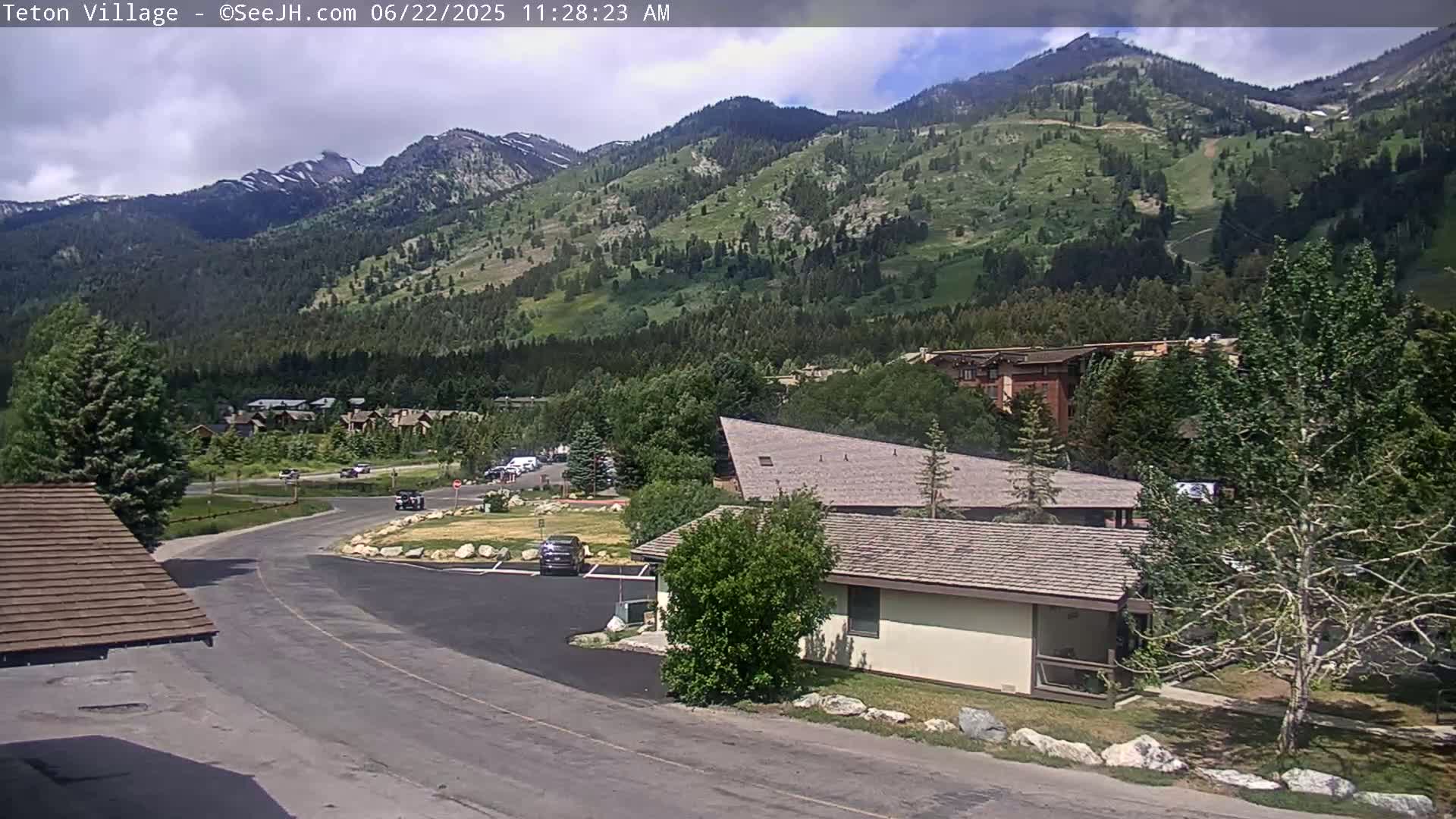 A parking lot and small buildings are situated in a valley, with a large mountain range covered in green vegetation visible in the background under partly cloudy skies.