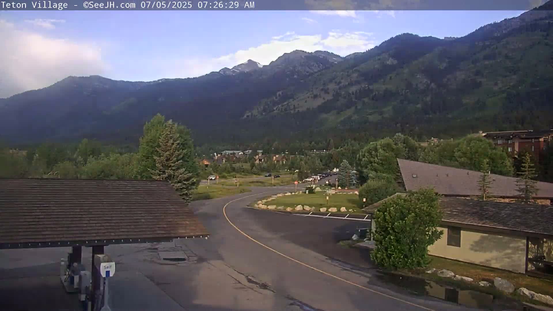 A mountain village with a gas station in the foreground, nestled in a valley under a partly cloudy sky.