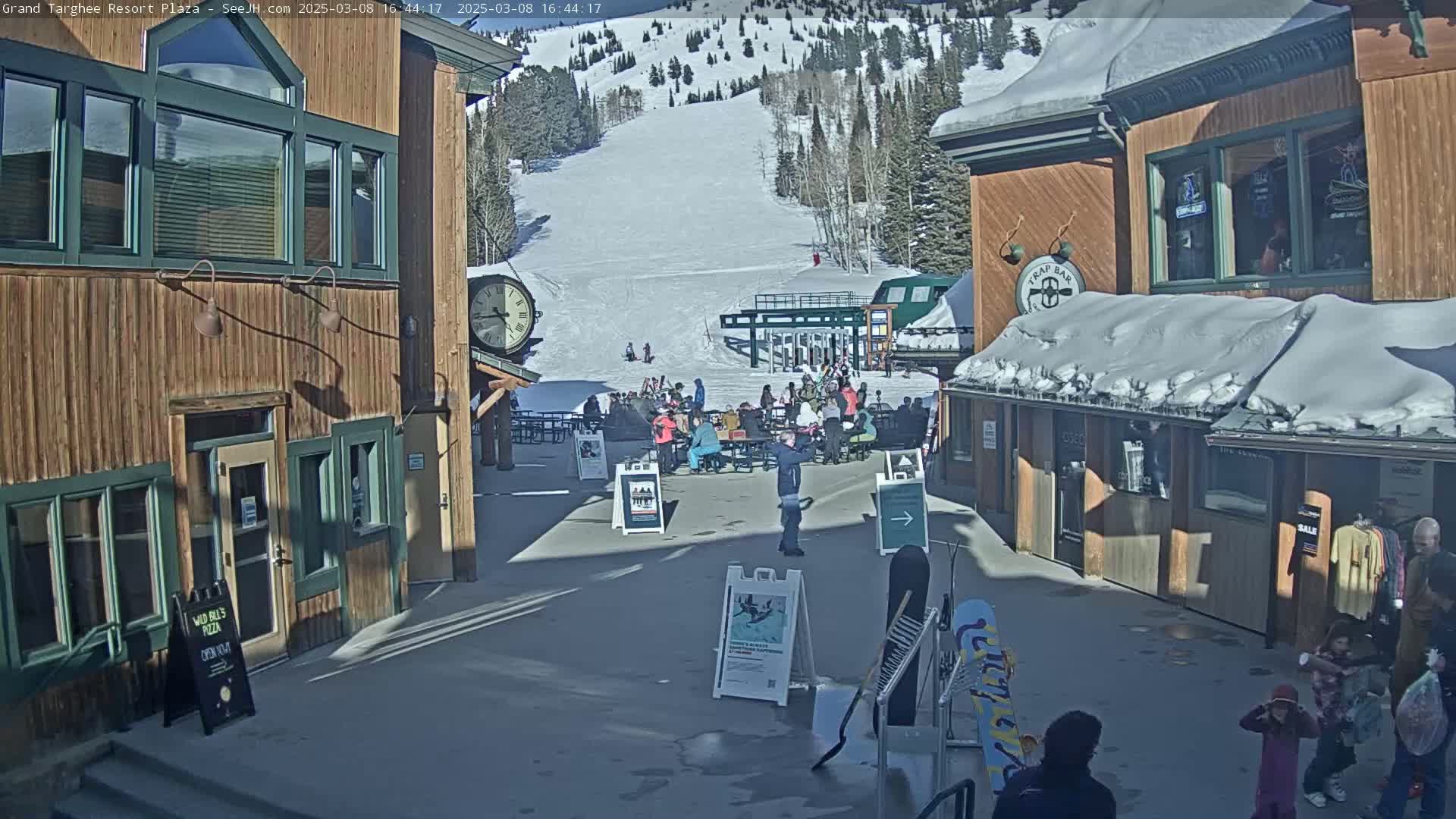 A sunny plaza with wooden buildings, a snow-covered ski slope in the background, and several people gathered in the plaza.