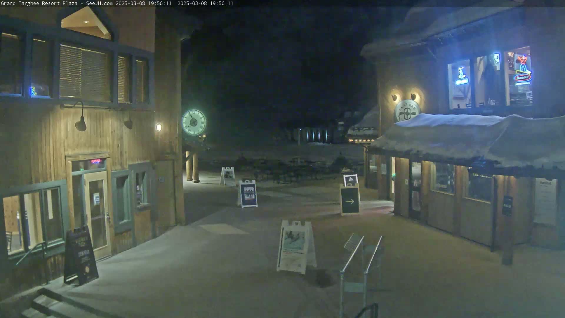 A snow-covered plaza at night, illuminated by lights on wooden buildings, features a clock and several signs.