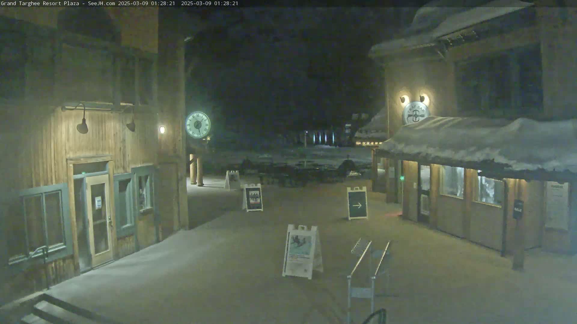 A snow-covered plaza at night is flanked by two wooden buildings, one with a clock, under dark, cloudy skies.