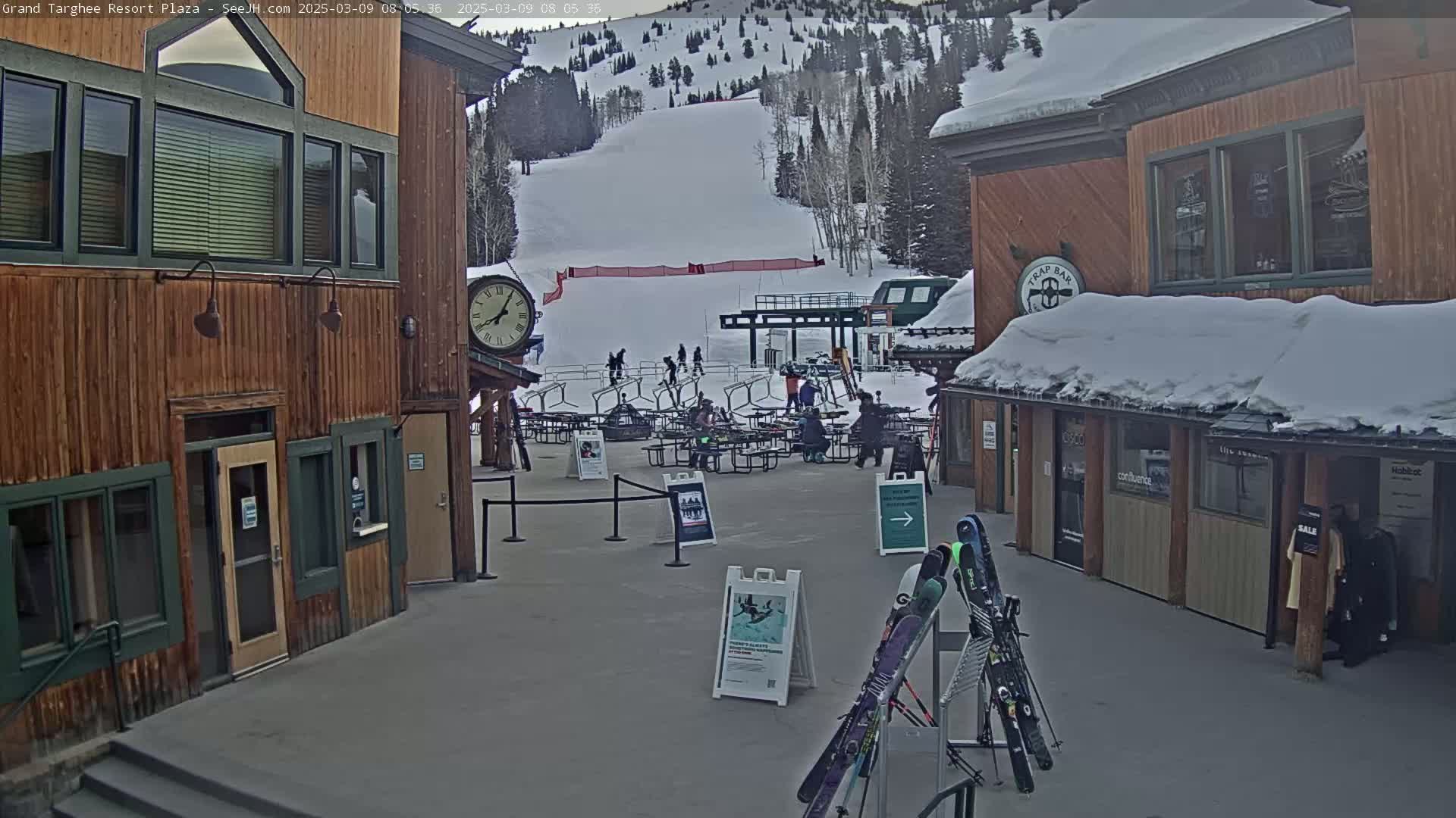 A plaza at a ski resort, covered in snow, shows a ski lift and slope in the background, with several people gathered around tables and chairs in the foreground.