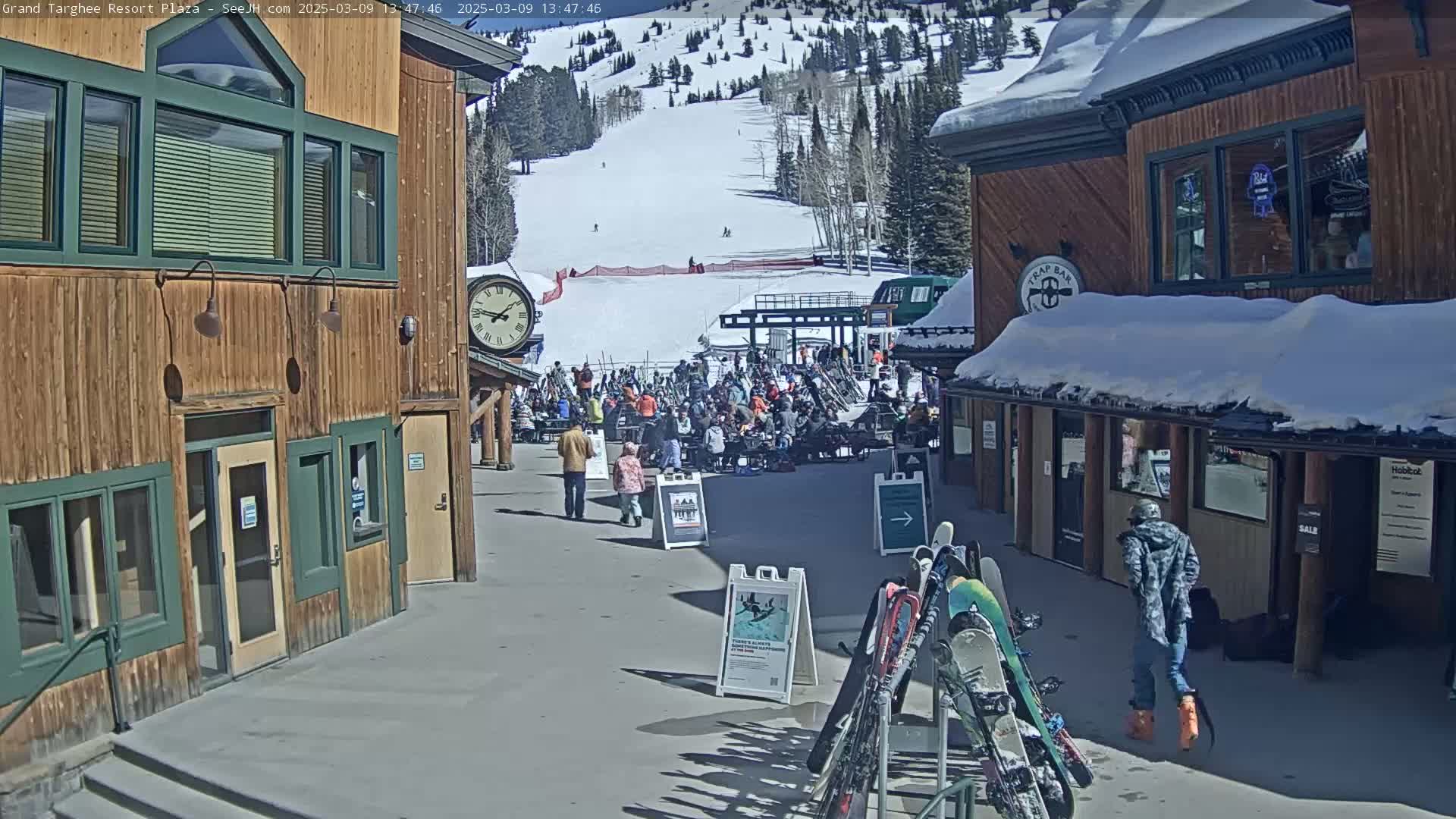 A sunny plaza at a ski resort with snow-covered buildings, a group of people gathered near a ski lift, and several pairs of skis leaning against a rack.