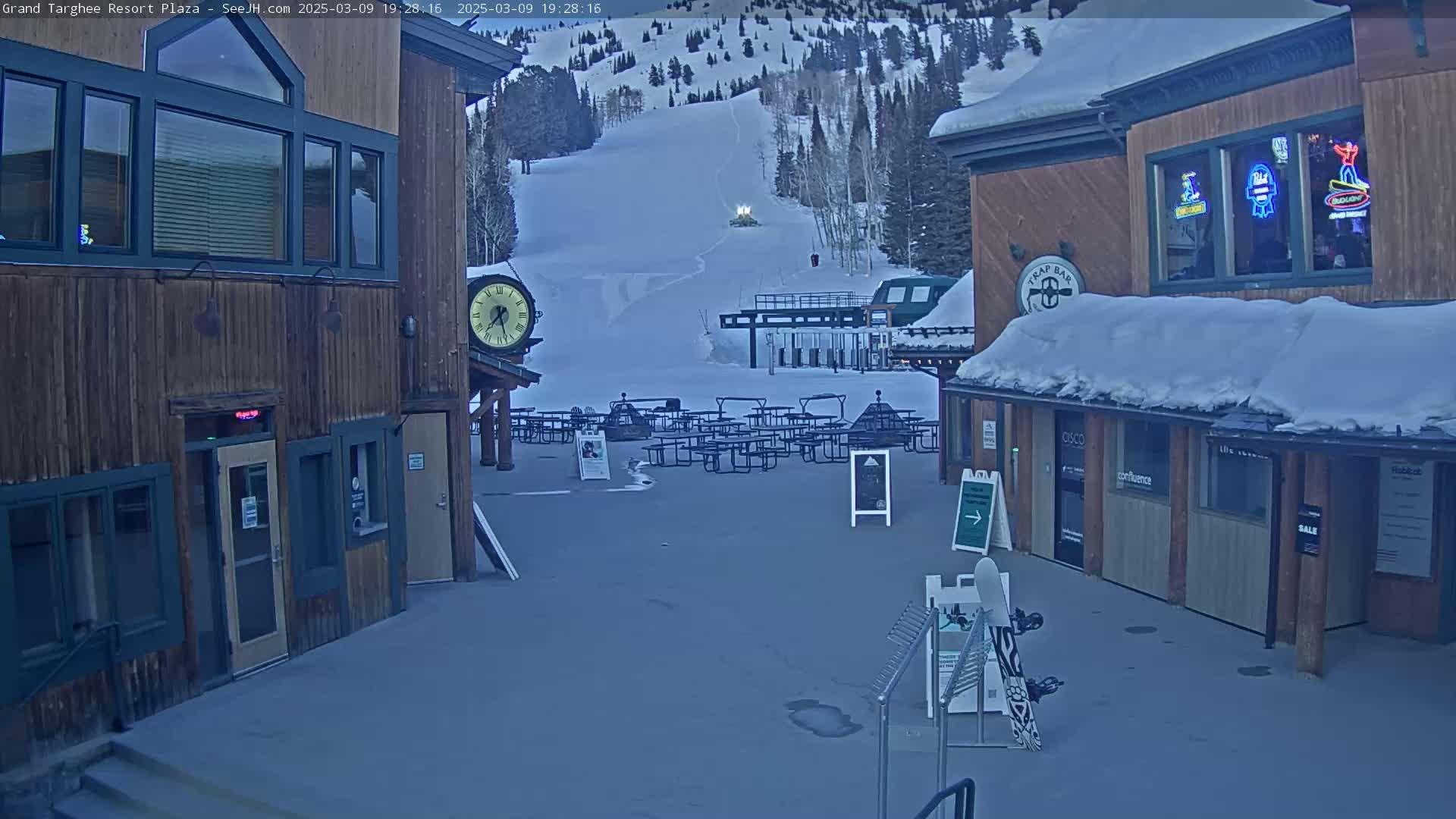 A snow-covered plaza with wooden buildings, outdoor seating, a ski lift in the background, and a mostly clear twilight sky.