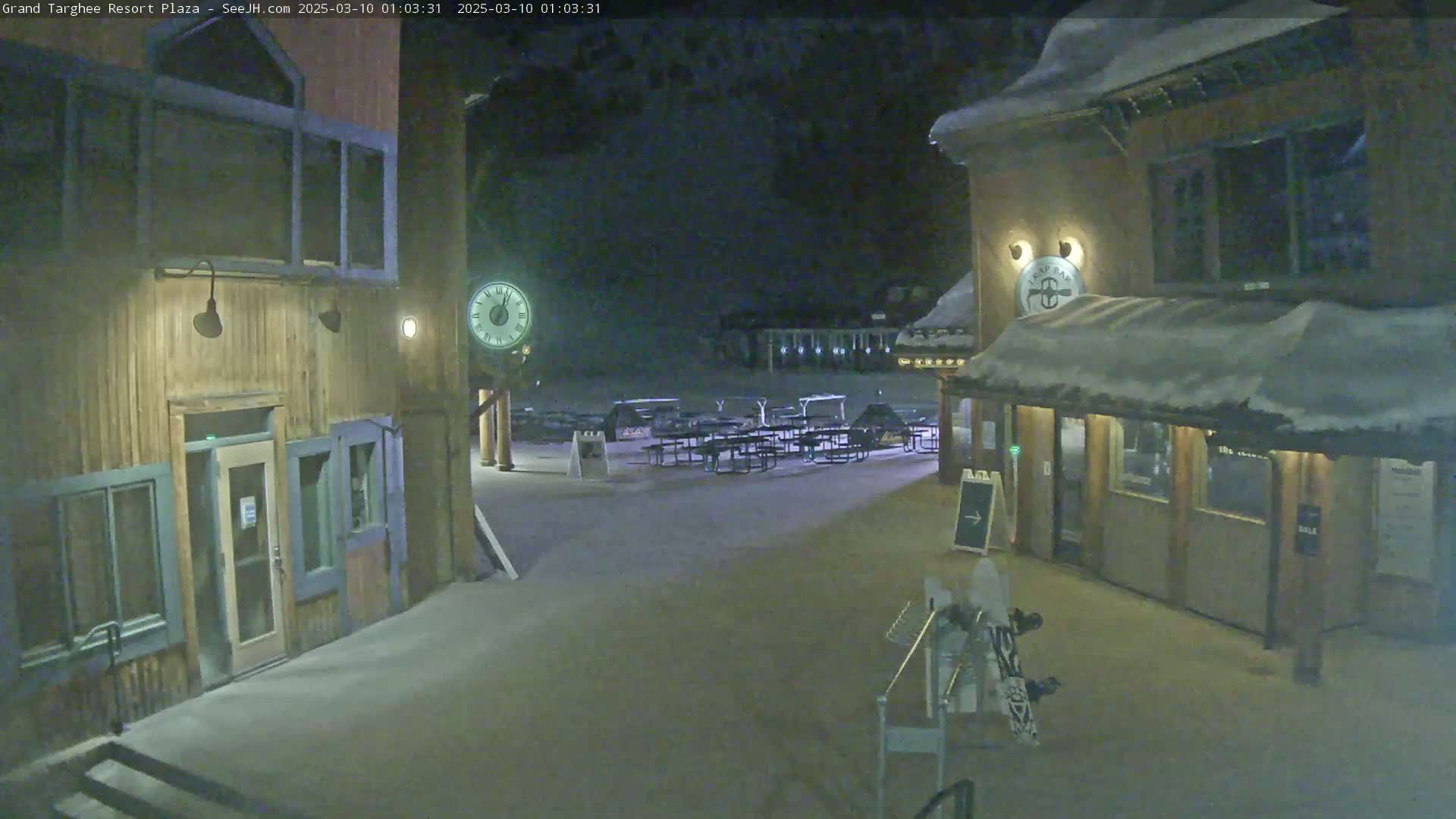 A nighttime snow-covered plaza with wooden buildings, picnic tables, and a clock.