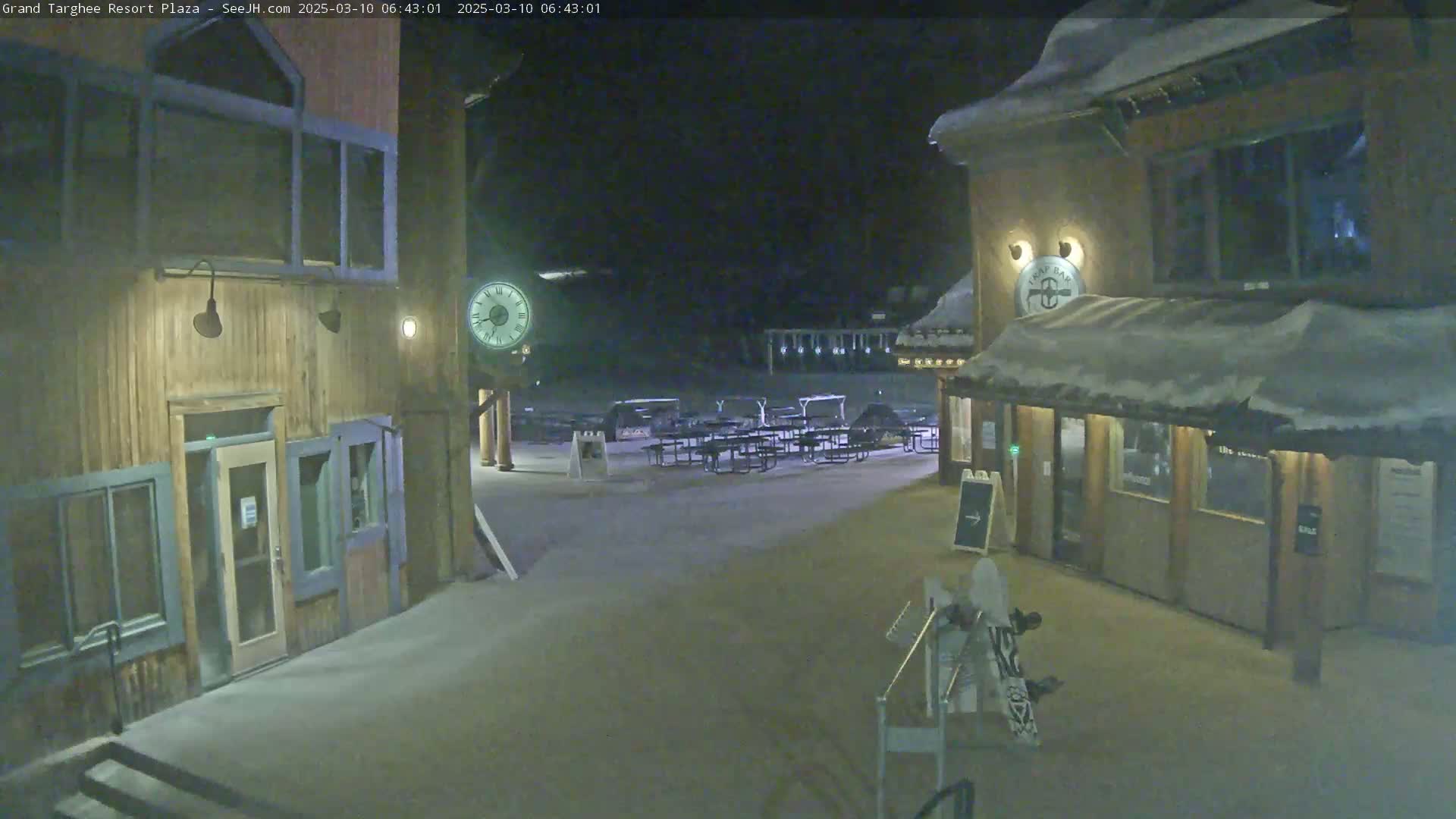 A snow-covered plaza at night, between two wooden buildings, with several empty picnic tables.