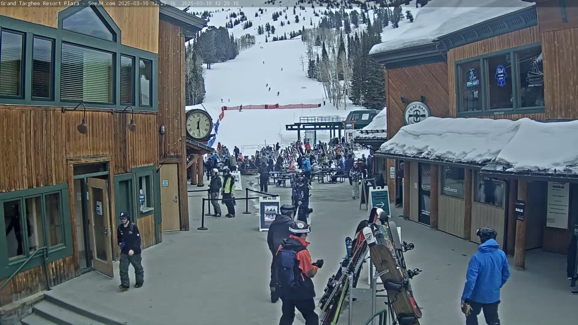 A plaza at a ski resort on a cloudy day, with snow-covered buildings and a crowd of people near ski racks, and a ski slope visible in the background.