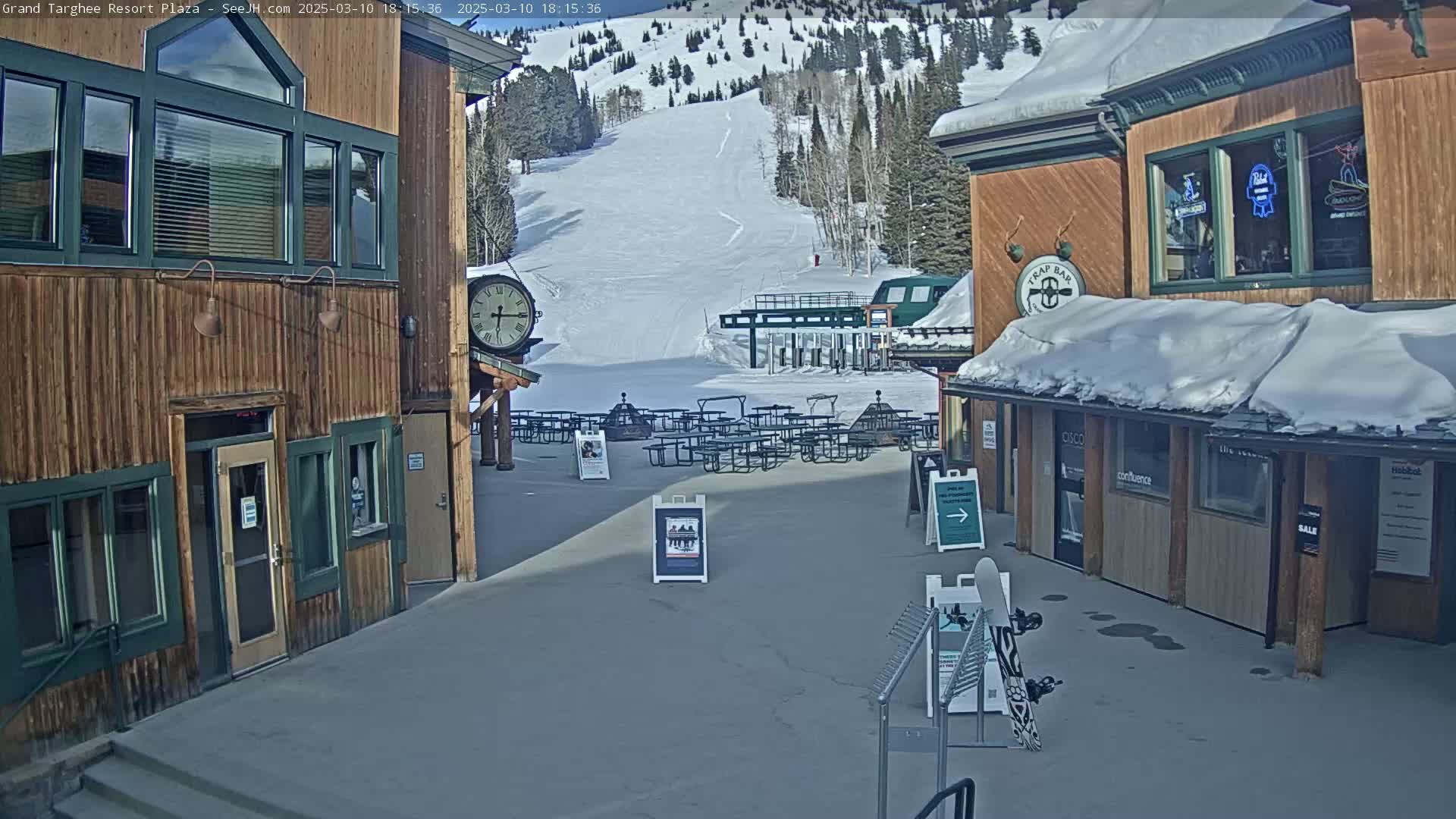 A snow-covered plaza with wooden buildings, picnic tables, and a ski slope in the background on a sunny day.