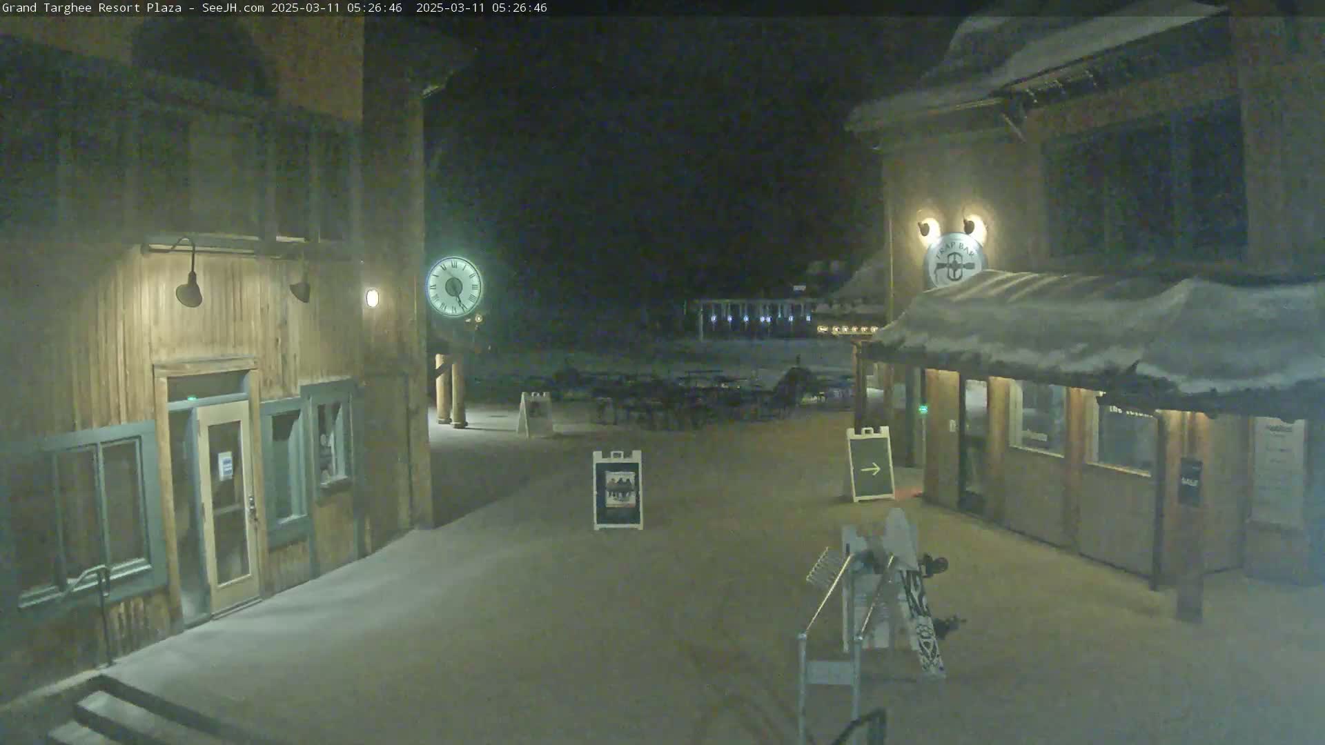 A snow-covered plaza at night, featuring two wooden buildings with outdoor lighting, and several empty tables and chairs.