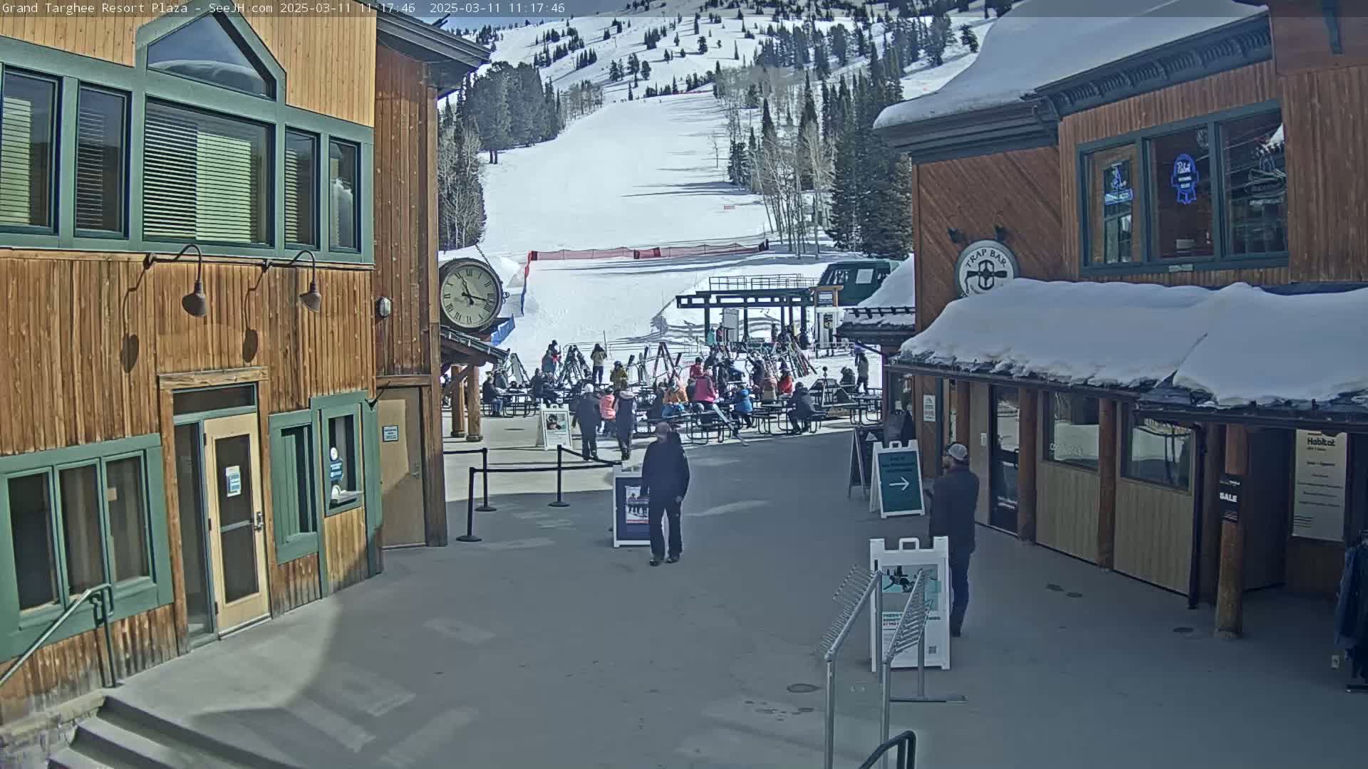 A plaza at a ski resort on a sunny day shows several people sitting at outdoor tables and others walking around, with a snow-covered mountainside visible in the background.