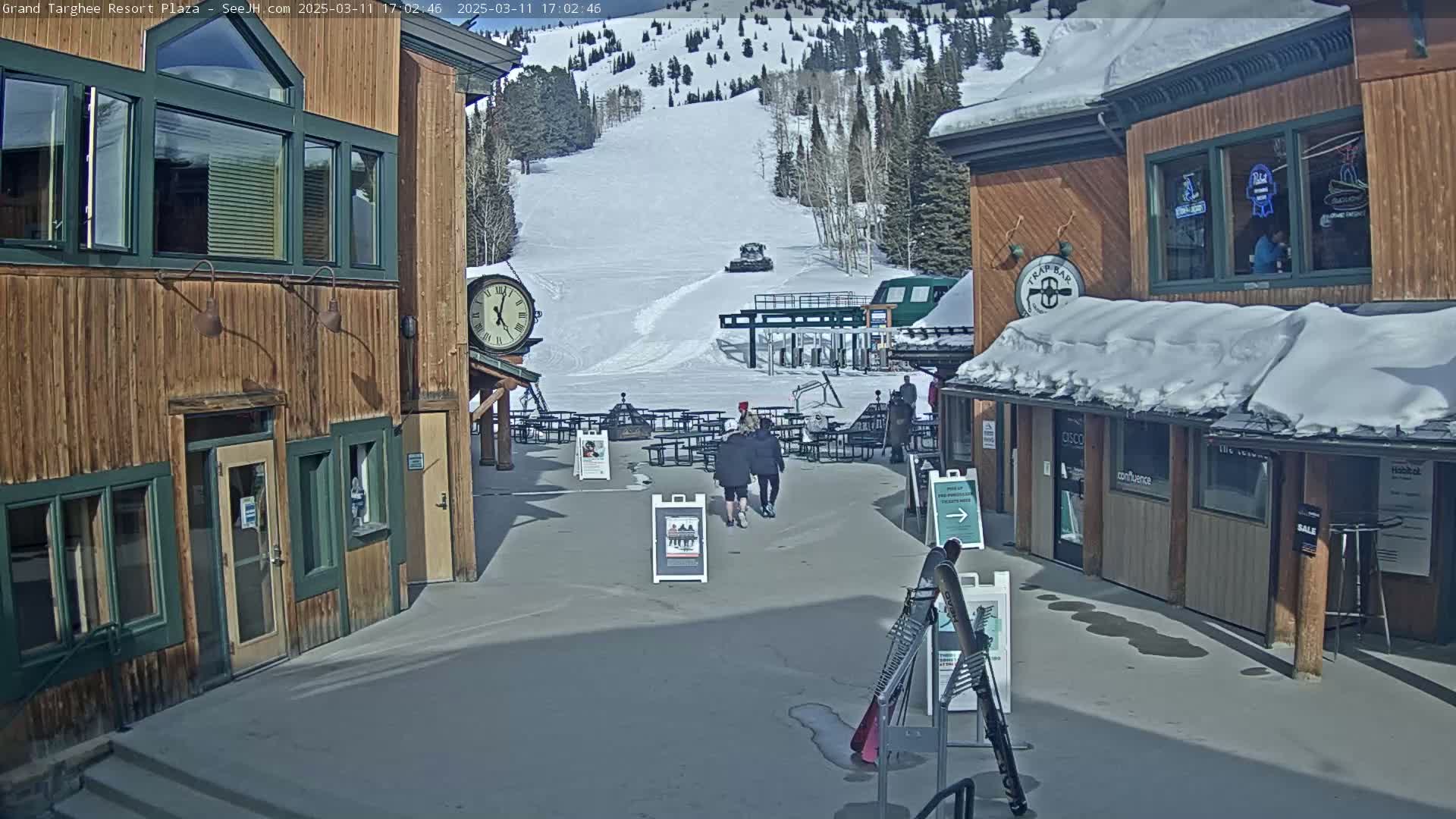 A sunny plaza with wooden buildings, snow-covered roofs, and a ski slope visible in the background.