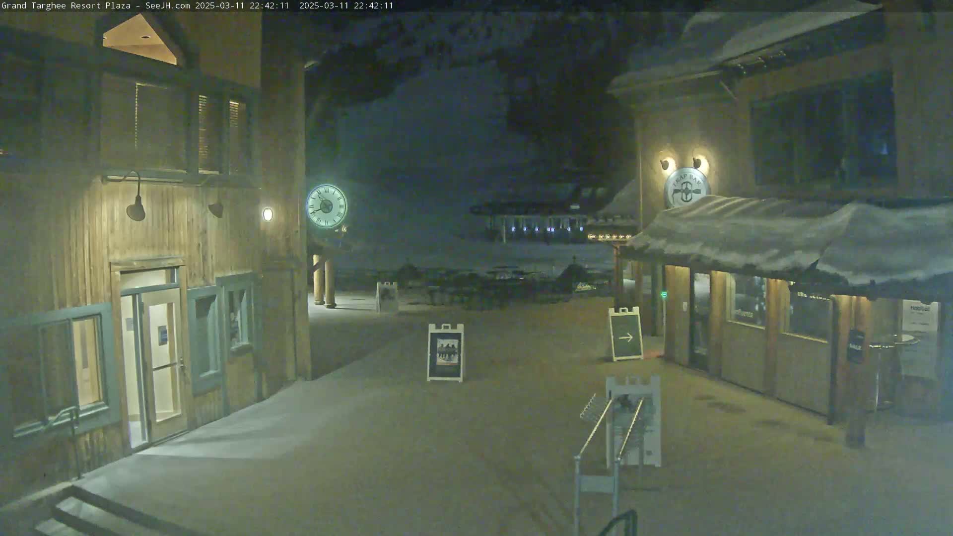 A snow-covered plaza at night is lit by building lights, featuring two wooden structures with entrances and a clock on a post.