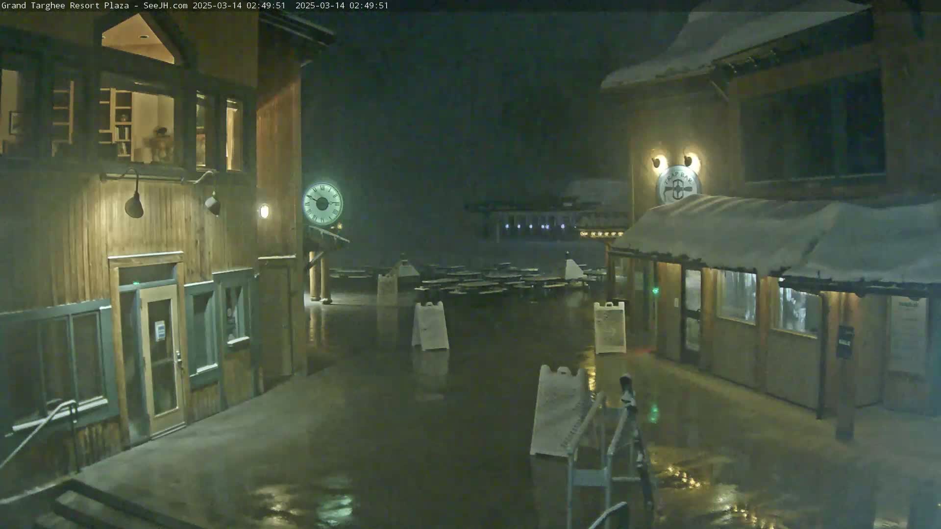 A nighttime view of a snow-covered plaza with two wooden buildings, one featuring a large clock, under a light snowfall.