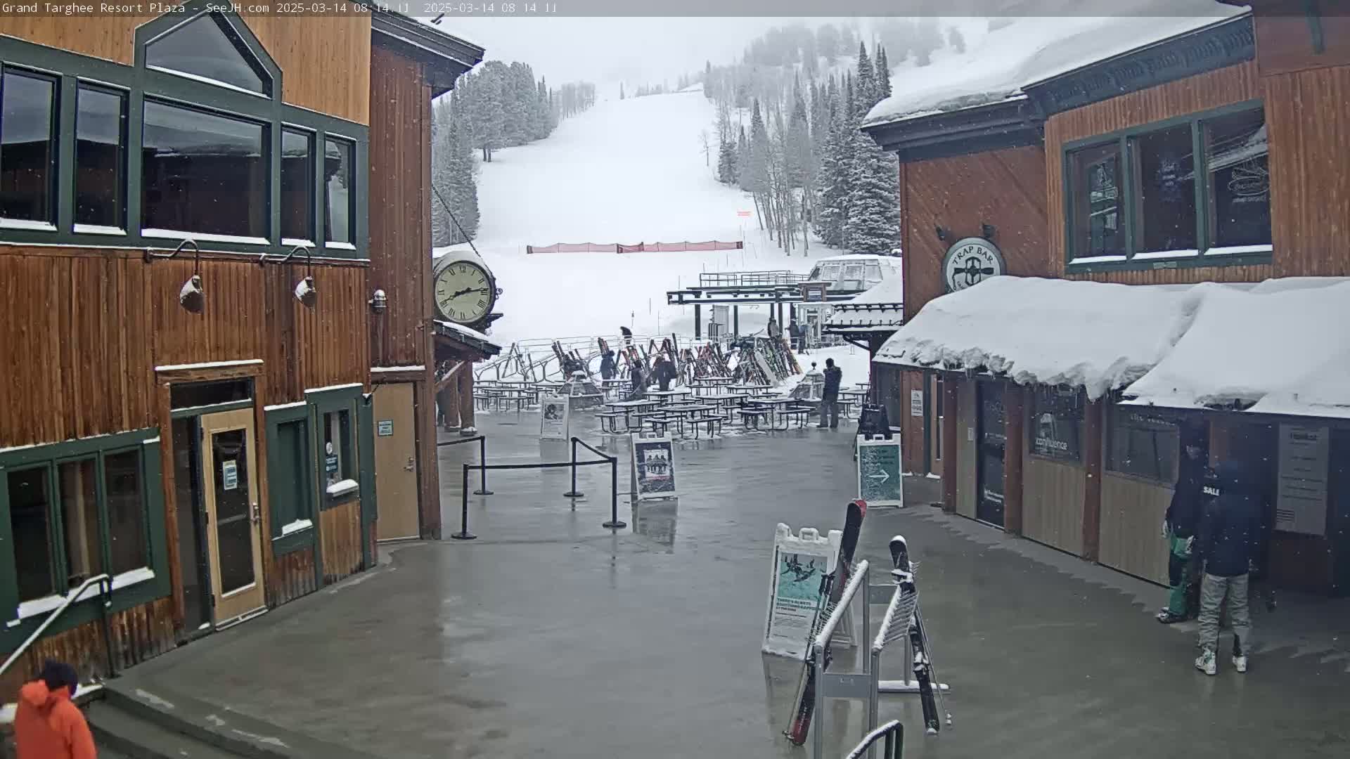 A plaza with wooden buildings, snow-covered picnic tables, and a ski slope in the background during a light snowfall.
