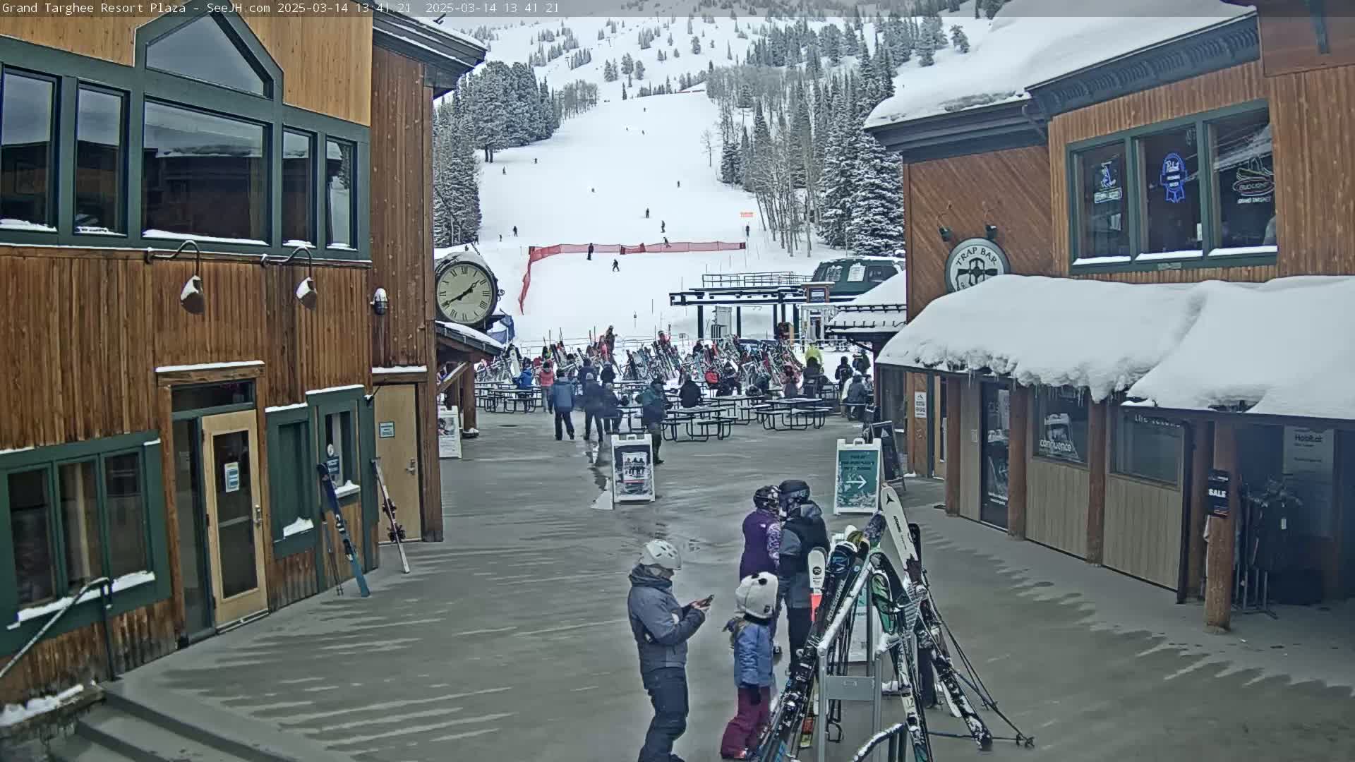 A snowy plaza scene shows people gathered around picnic tables near ski racks, with a ski slope visible in the background and snow-covered buildings on either side.