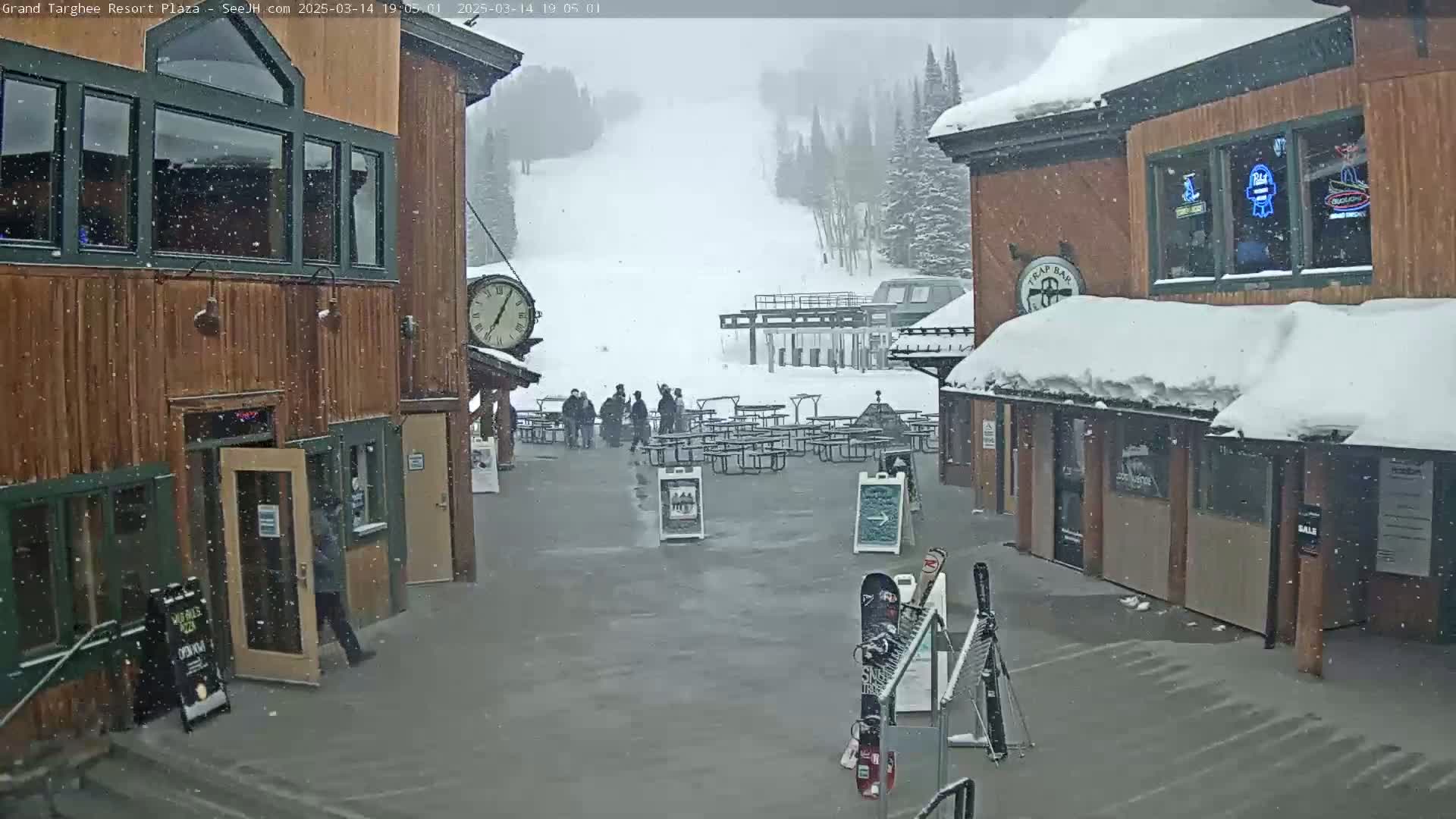 Snow falls on a plaza with wooden buildings, outdoor seating, and a ski slope in the background.