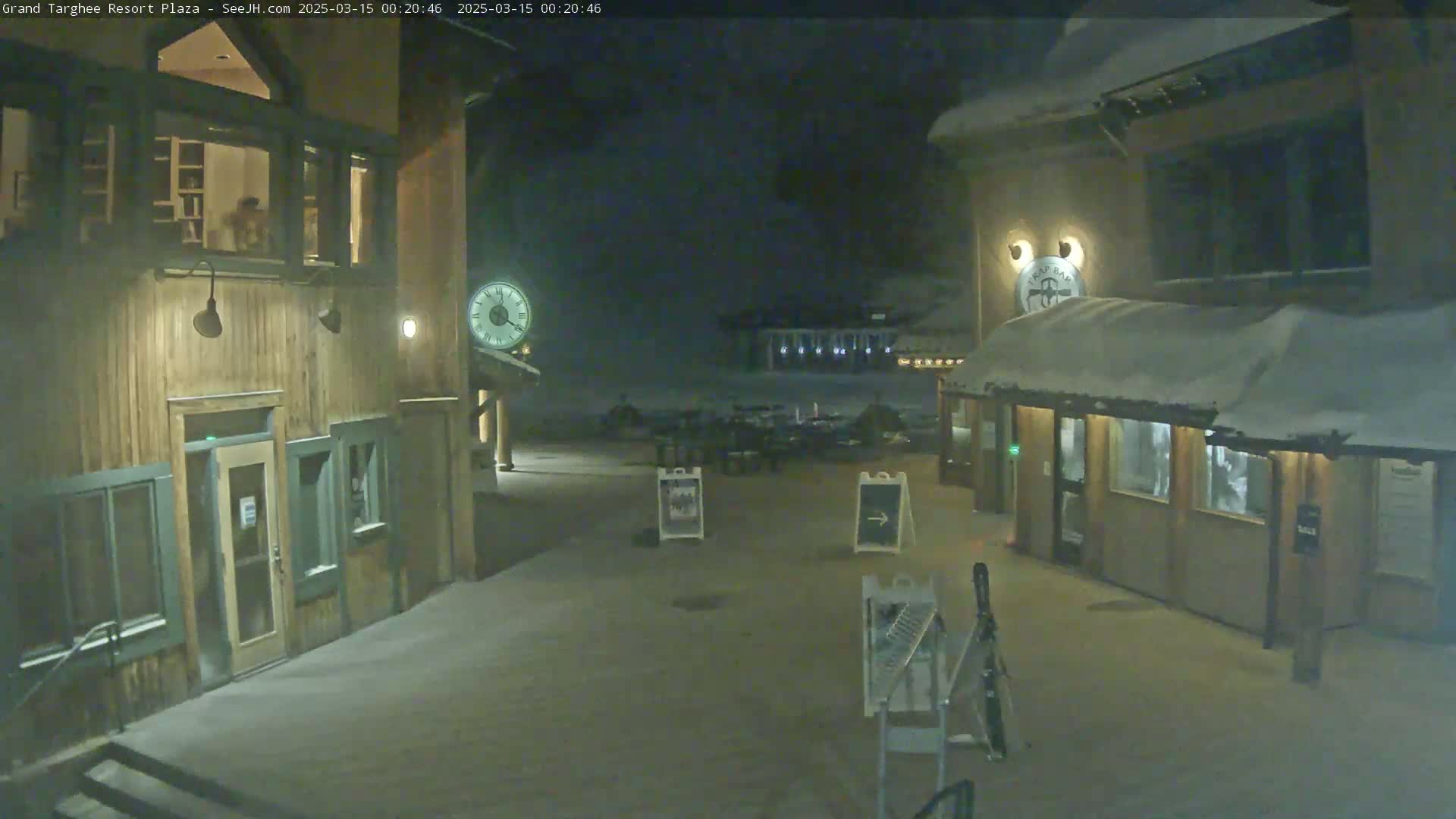 A snow-covered plaza at night, illuminated by building lights, shows several wooden buildings with large windows.