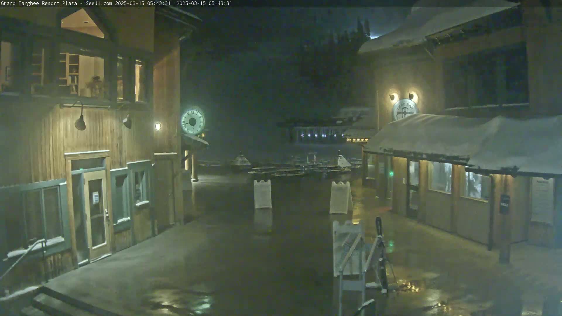 A snow-covered plaza at night, illuminated by dim lights, shows several wooden buildings with reflective, wet ground.