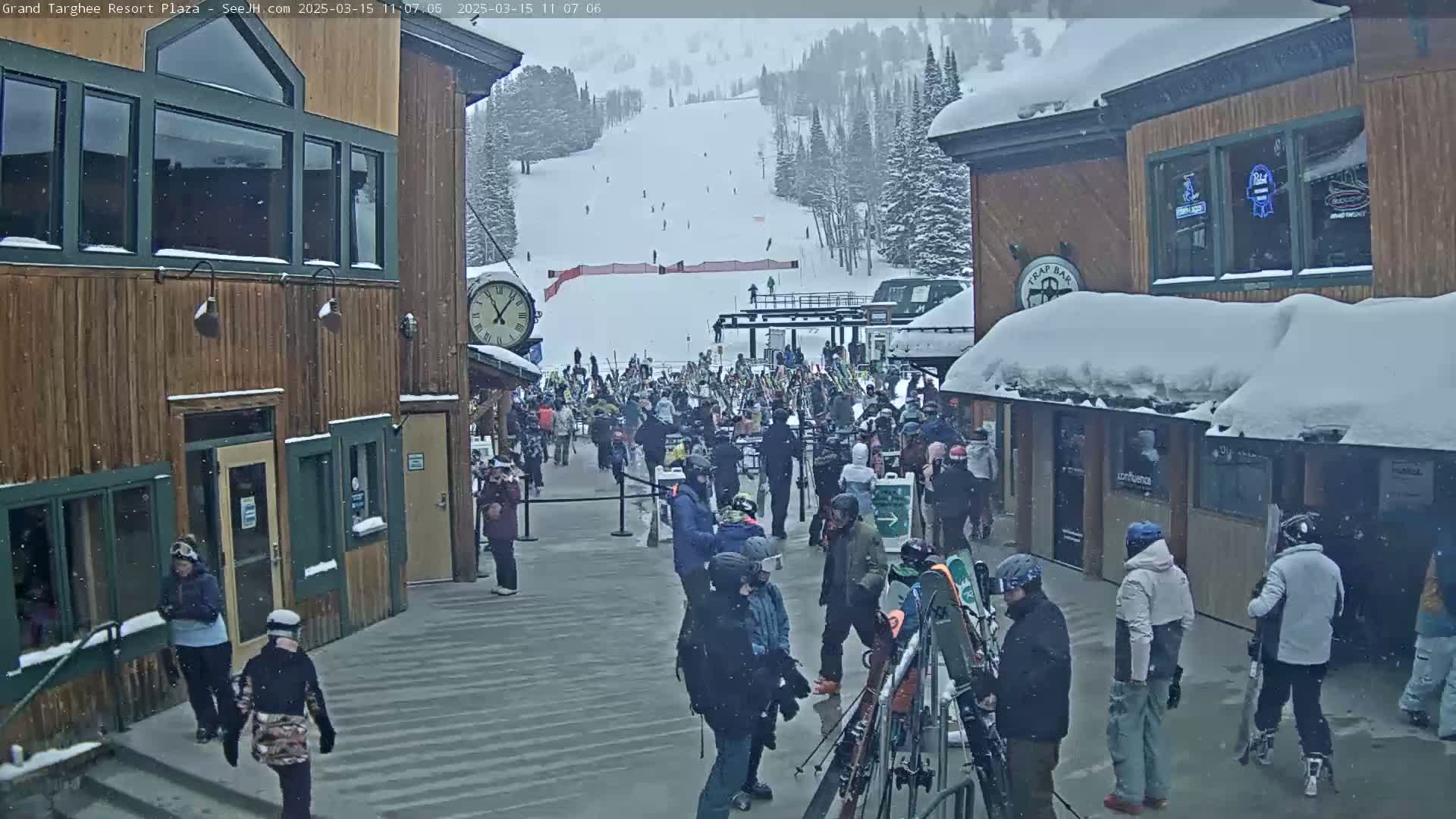 A snowy scene shows a crowd of people gathered outside wooden buildings near a ski slope.