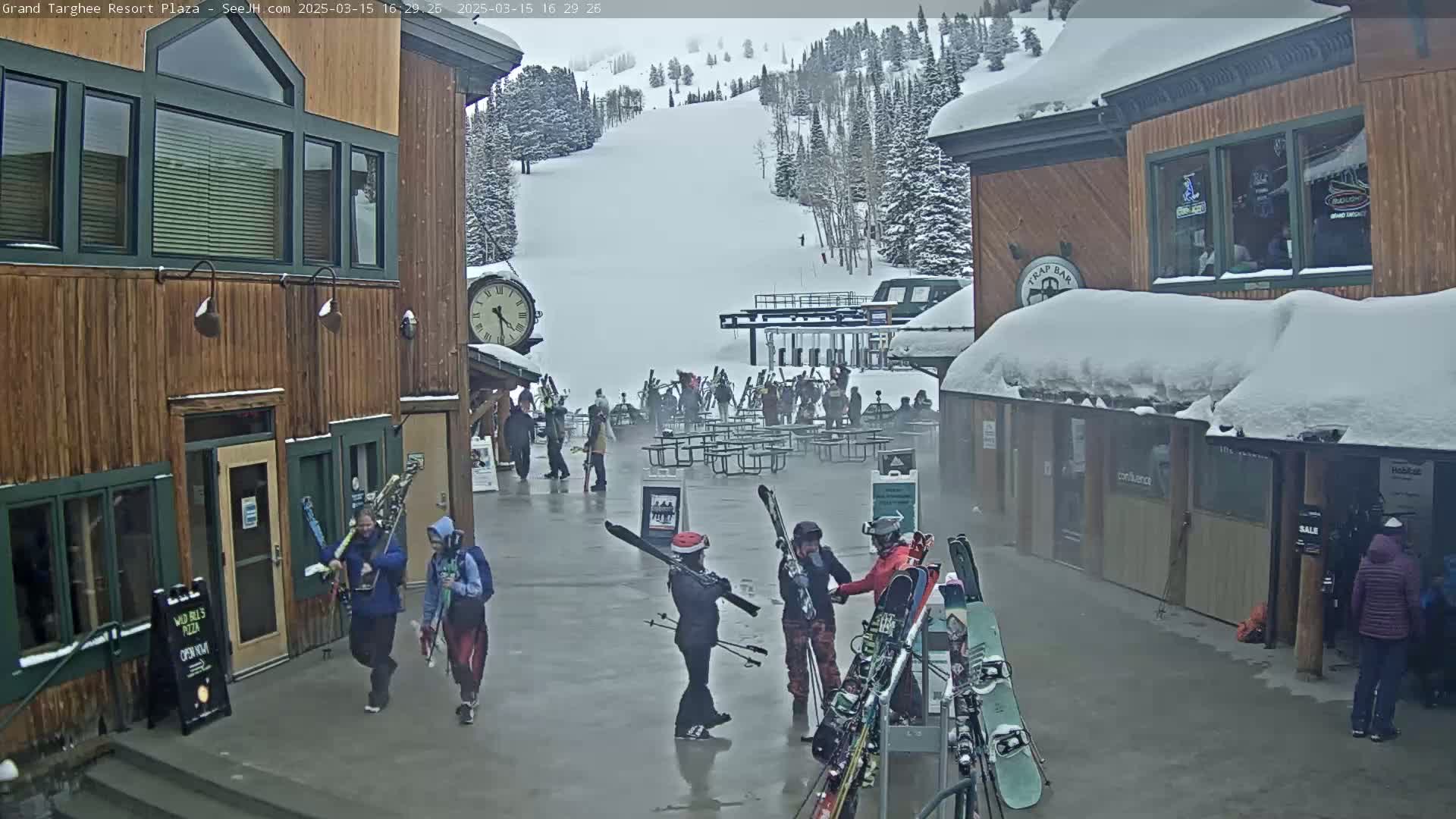 A snowy plaza at a ski resort with several buildings, people carrying skis, and a ski lift visible in the background under overcast skies.