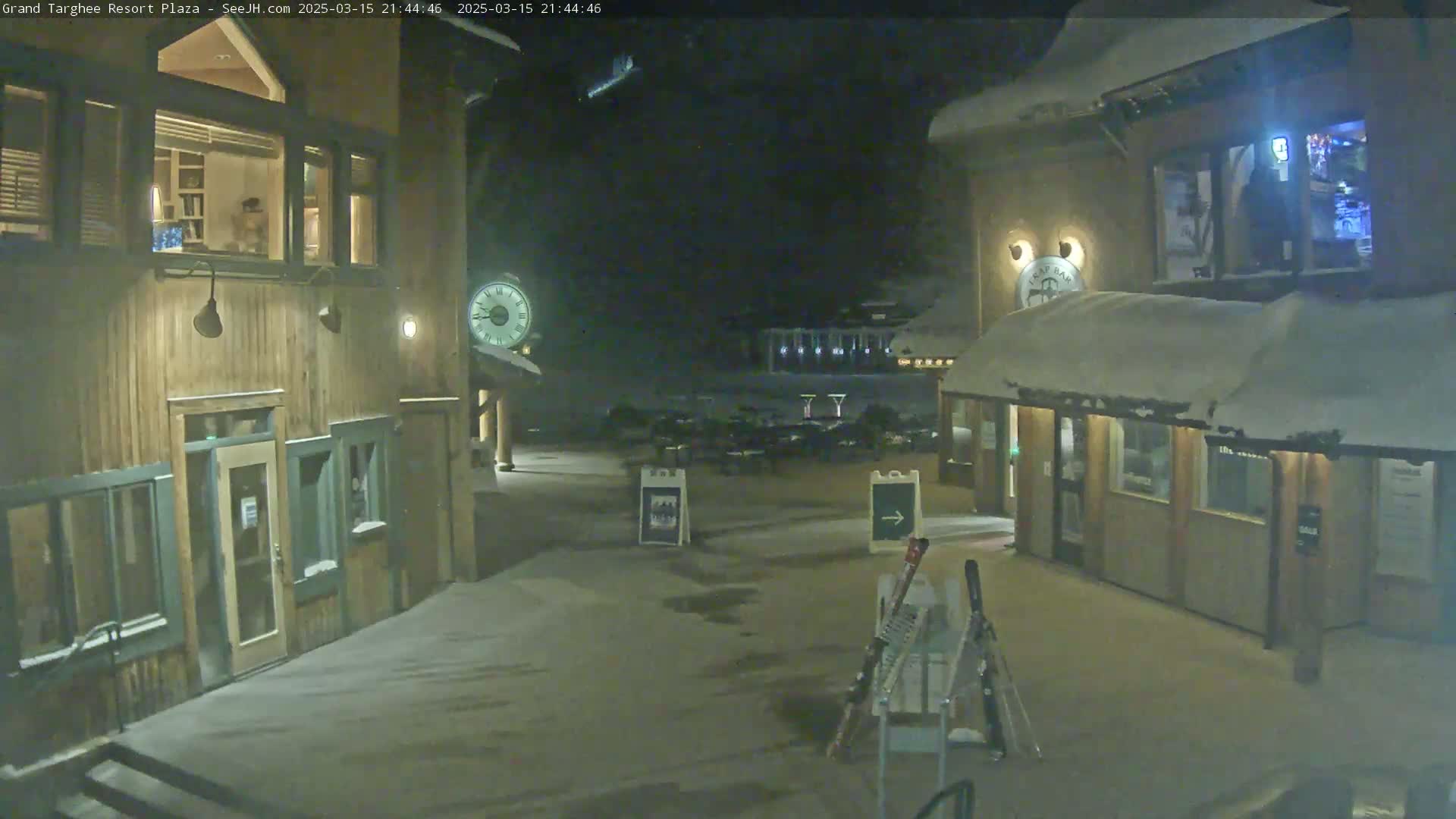 A snow-covered plaza at night, illuminated by lights, shows two wood buildings with lit windows and a clock on one, skis leaning against a stand in the center, and a mostly empty parking lot in the background.
