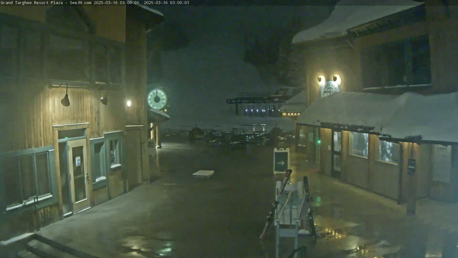 A snow-covered plaza at night, between two wooden buildings, is lightly illuminated with some puddles reflecting light on the wet ground.