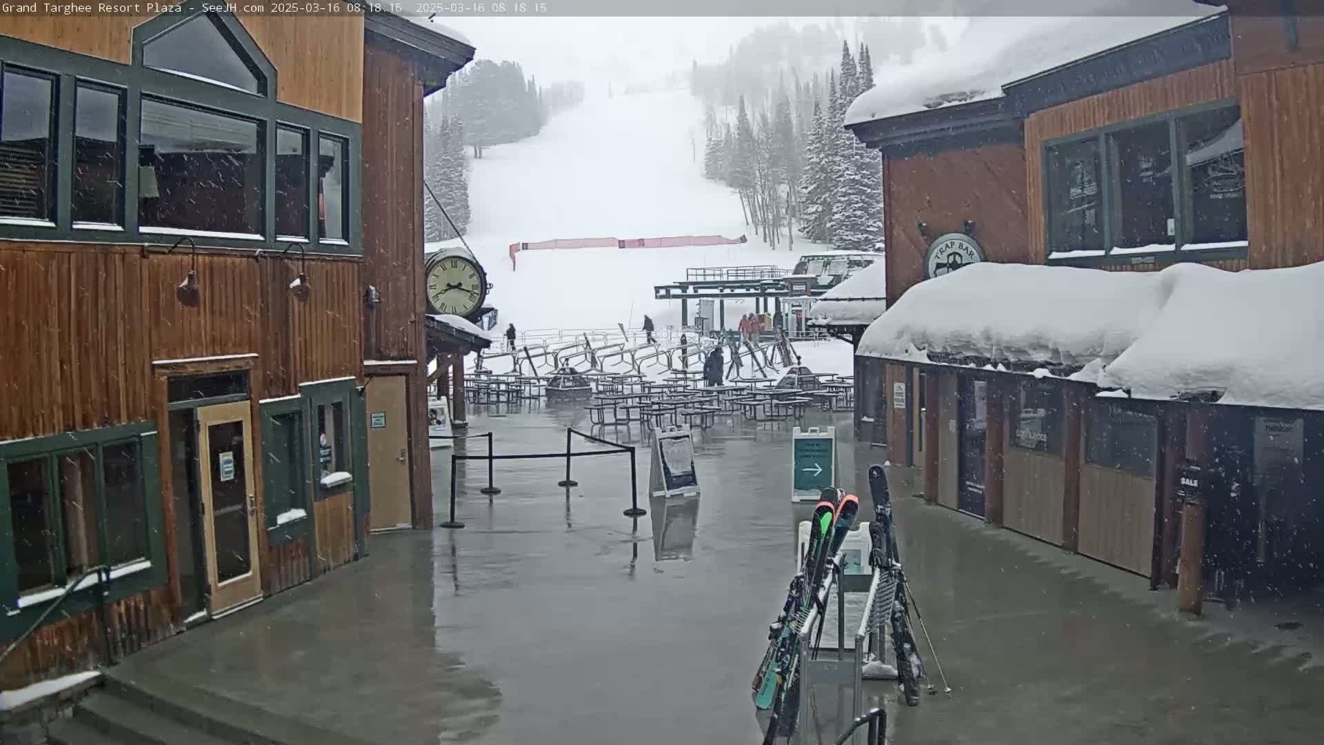 A snow-covered plaza at a ski resort, with wet pavement, is seen during a light snowfall, featuring several buildings, outdoor seating, a ski lift, and a clock.