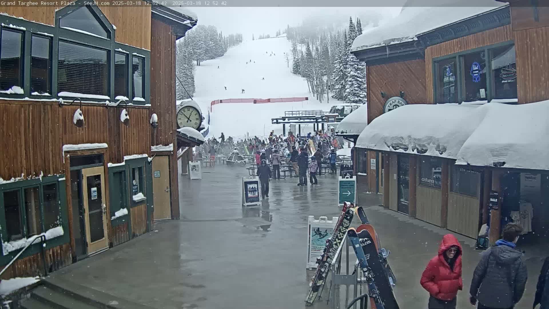 A snowy plaza with wooden buildings, a ski slope in the background, and several people milling about in light snow.