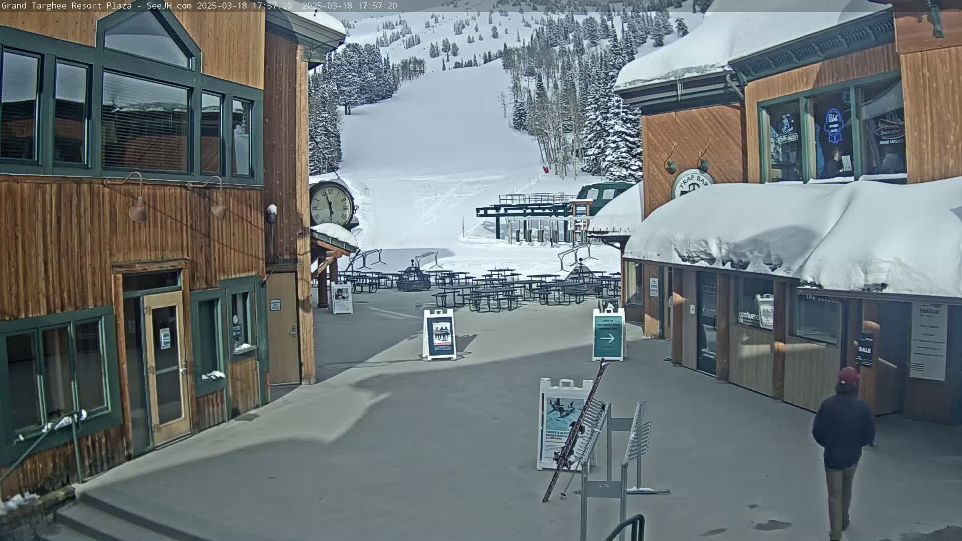 A plaza with wooden buildings, snow-covered roofs, and picnic tables overlooks a snow-covered ski slope on a sunny day.