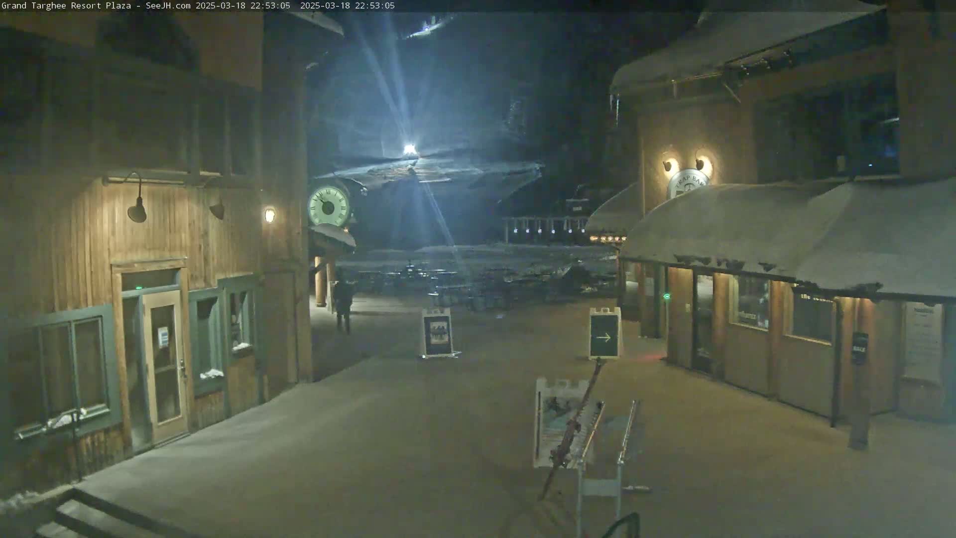 A snow-covered plaza at night, illuminated by lights, with wooden buildings and a person walking in the distance.