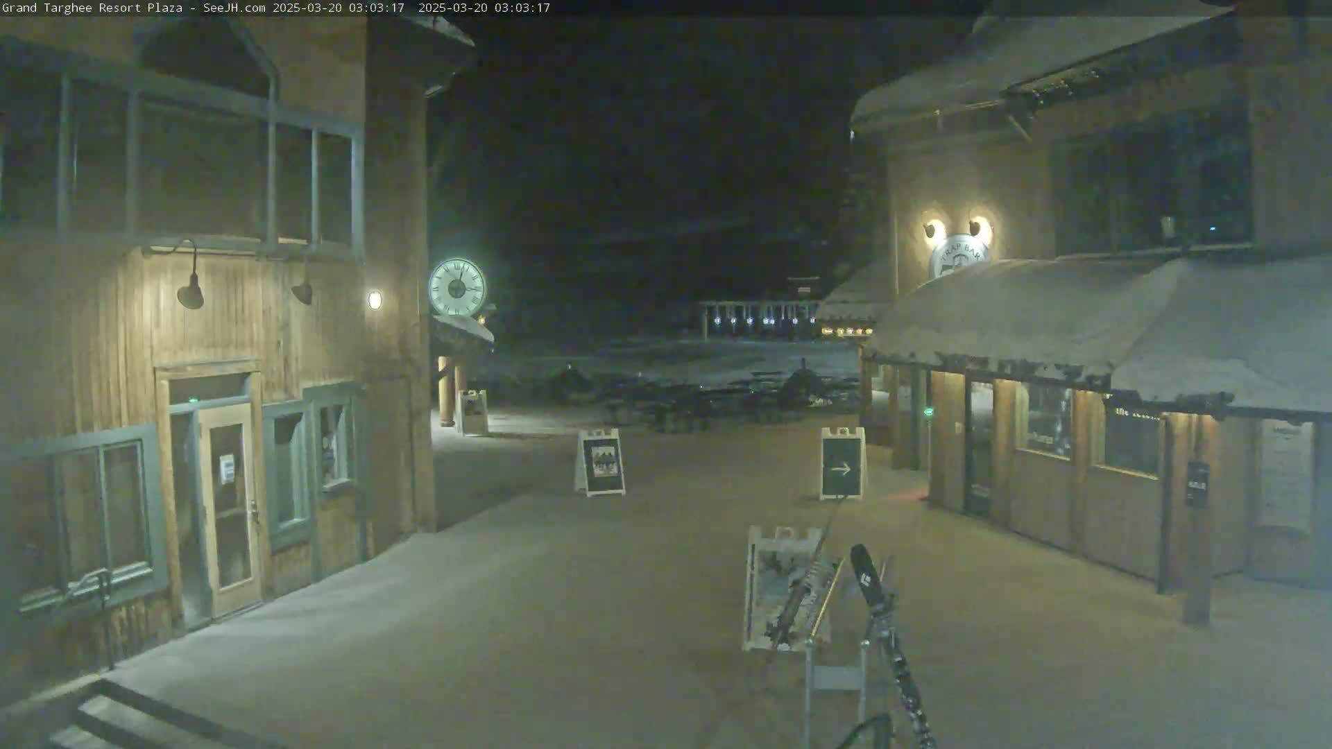 A snow-covered plaza at night is lit by several lamps, showing two wooden buildings with clocks and signage.