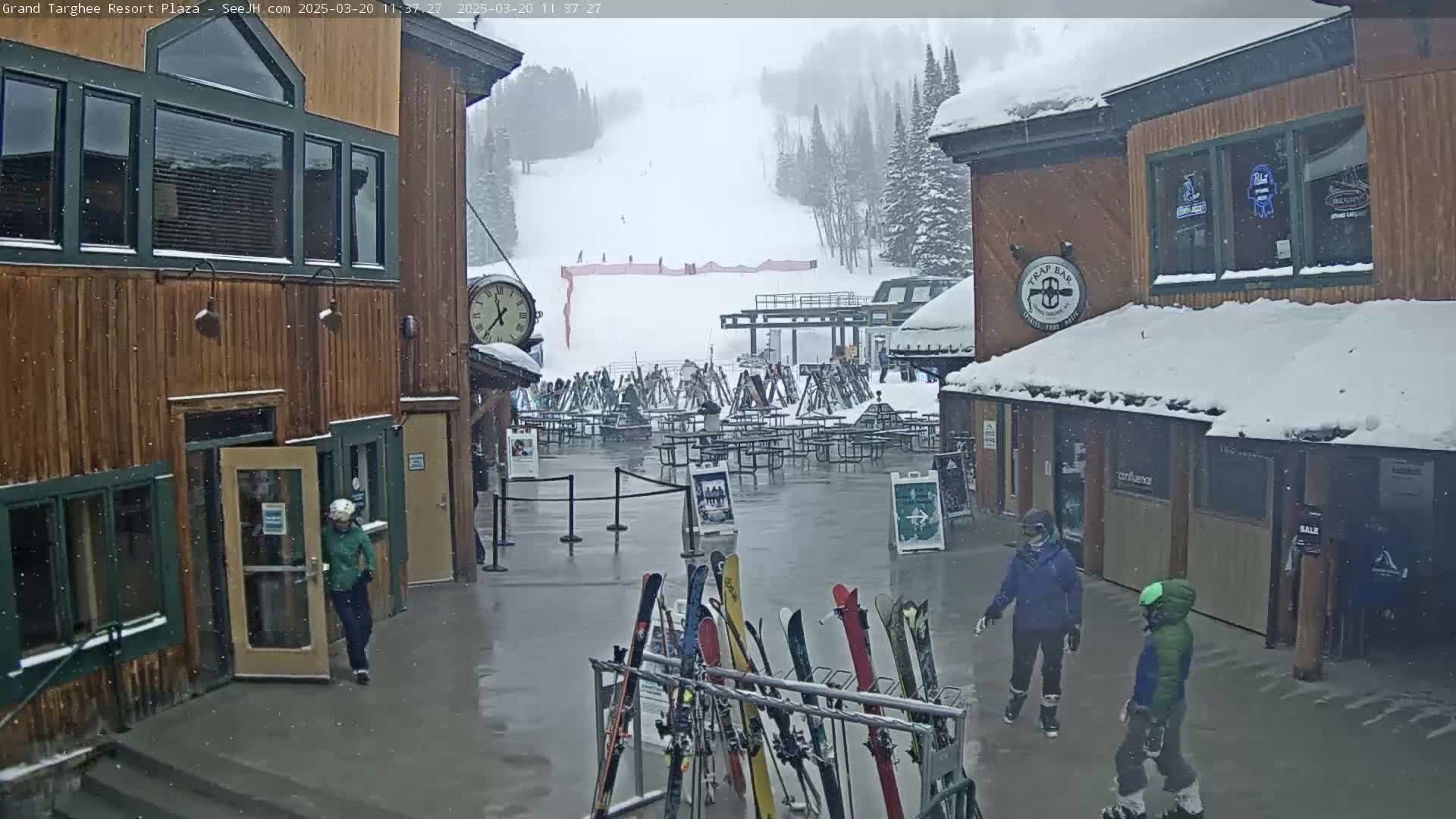 A snow-covered plaza at a ski resort, showing several people walking amidst a light snowfall, with a ski lift and slope visible in the background.